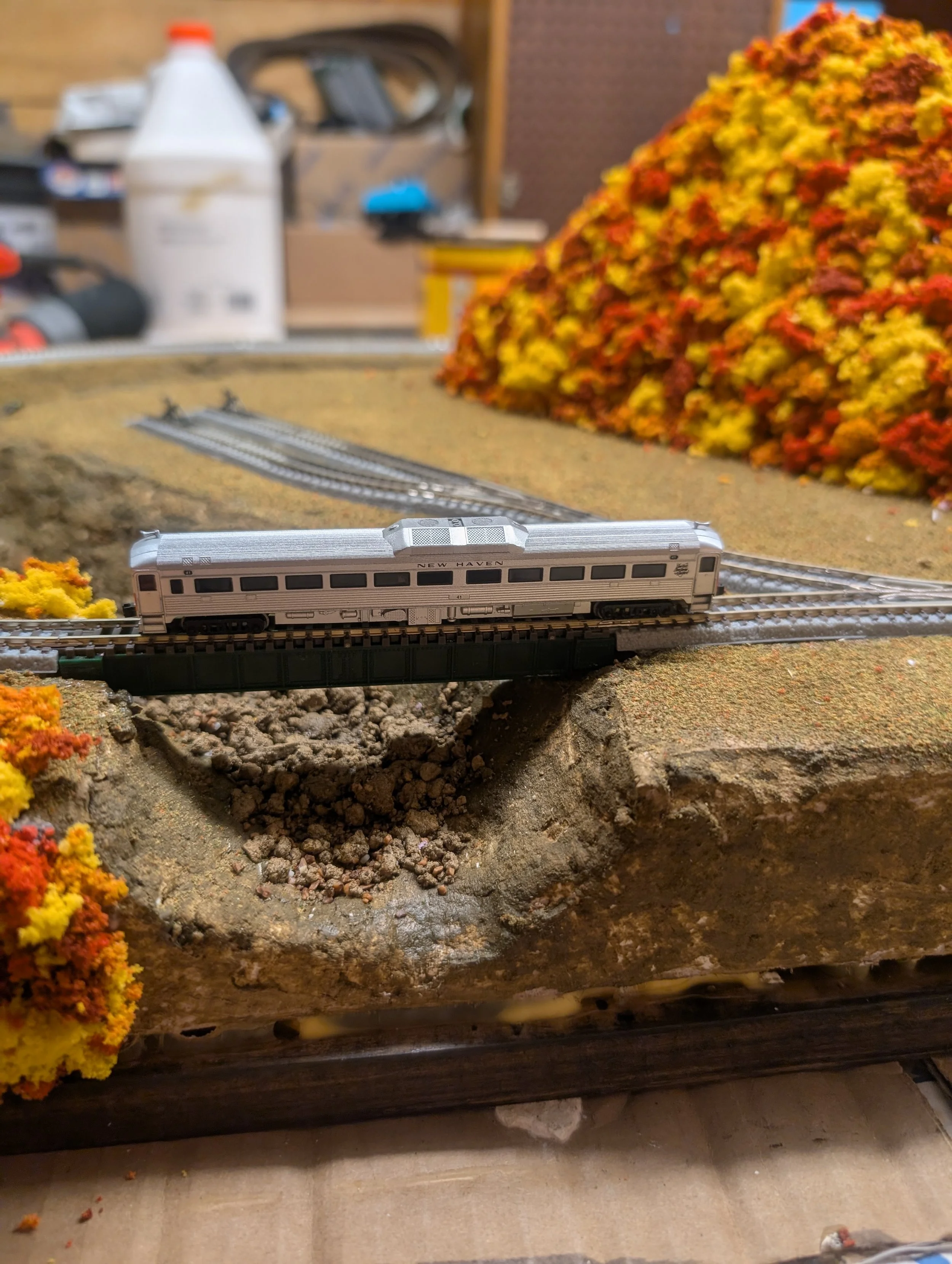 A miniature model train on tracks with a landscape featuring rocks and colorful foliage.