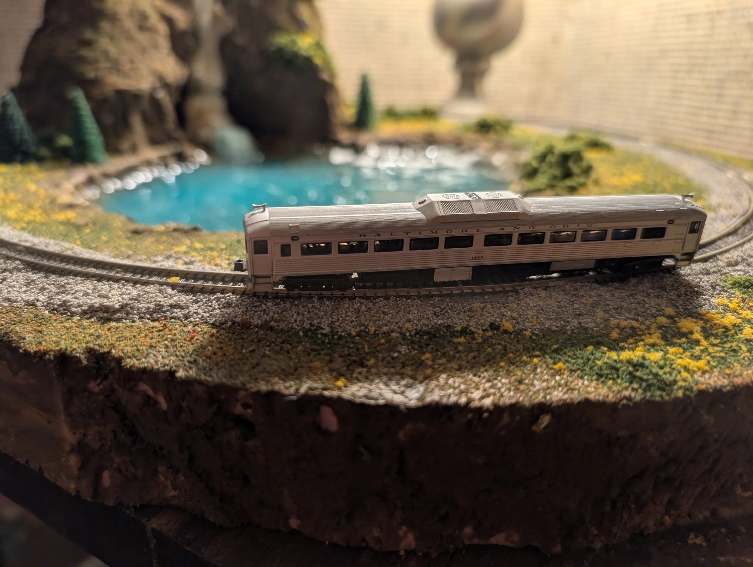 A detailed model train set featuring a silver passenger train on curved tracks, with a scenic landscape including a pond, trees, and a brick wall in the background.