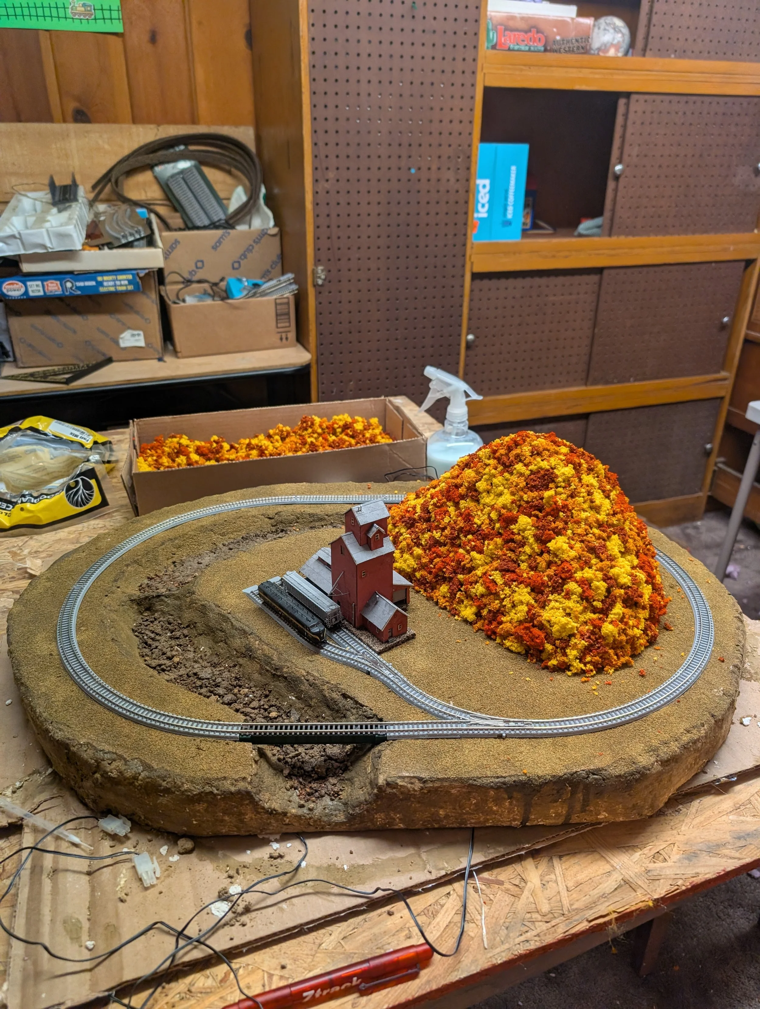 Model train setup on a wooden table with a small train, a red barn, a large yellow-orange tree, and a landscape base. In the background, shelves with boxes, a spray bottle, and miscellaneous tools and supplies.