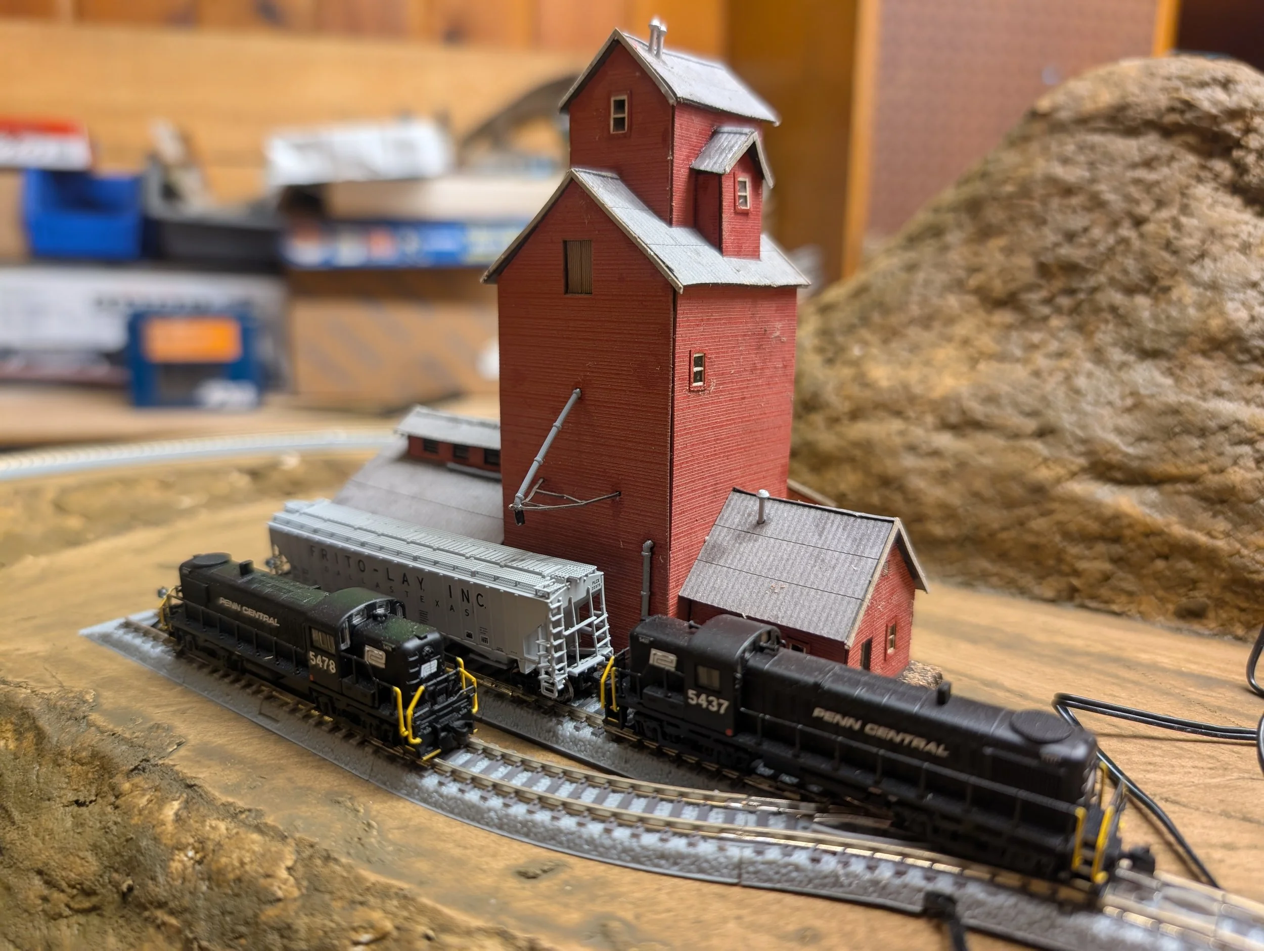 Model train set with a black train on curved tracks next to a red silo and small red building, with a large rock in the background.