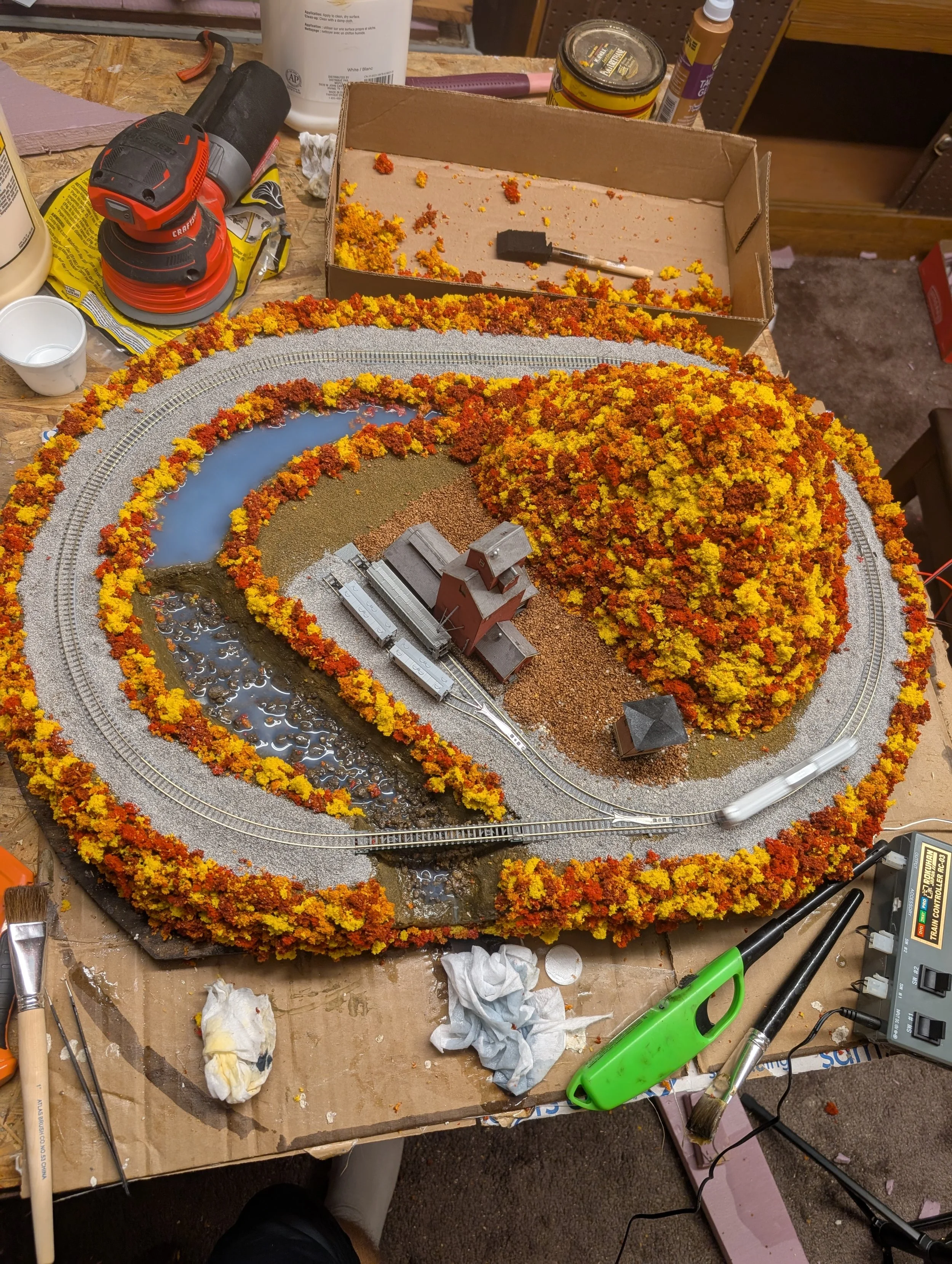A detailed model train set surrounded by artificial trees with fall colors, water, and mountains, set on a worktable with tools and craft supplies around it.