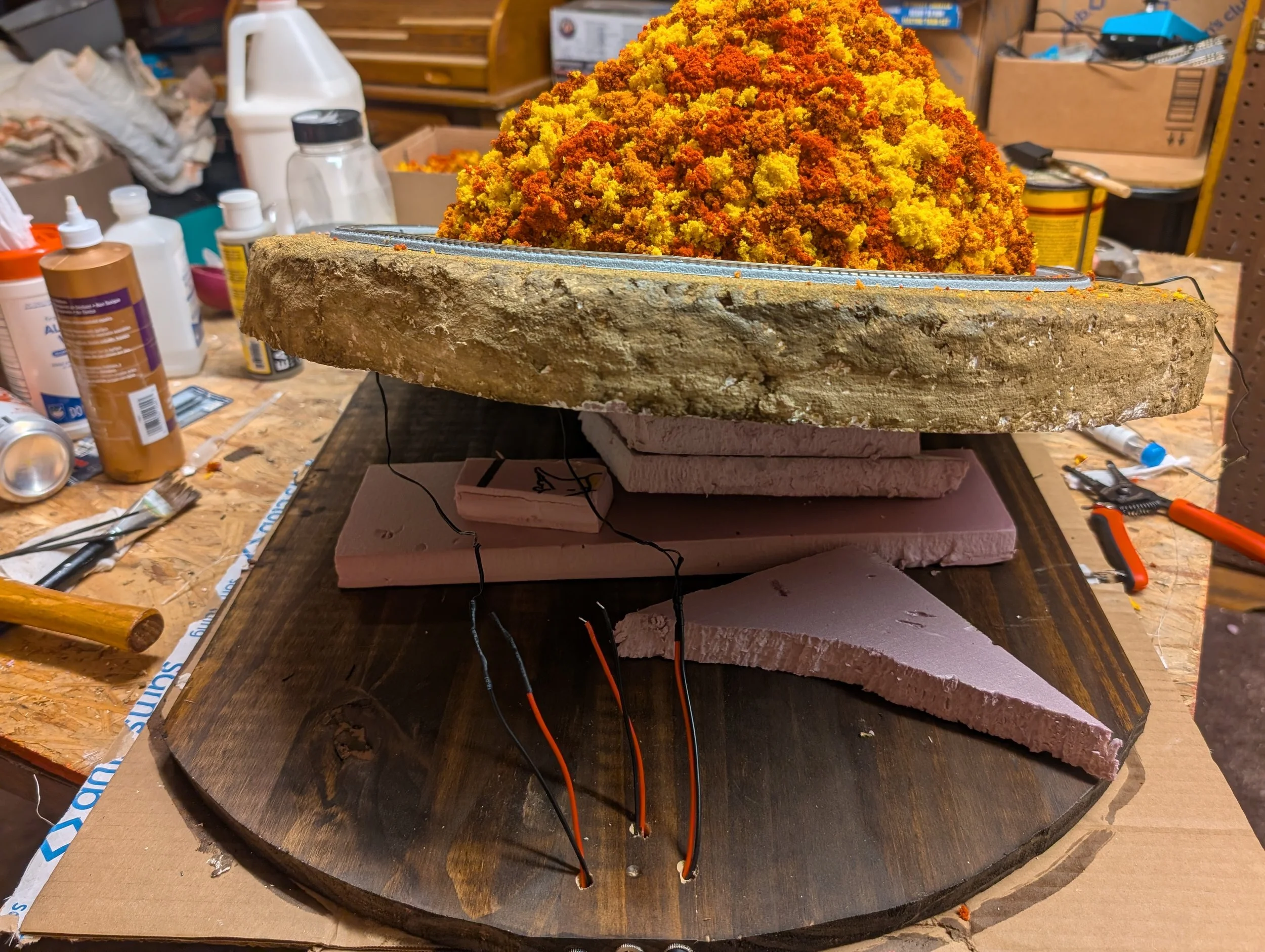 A model train set in progress on a workbench, featuring a mountainous landscape made of foam and a hill covered with orange and yellow foliage, with wires and tools around.