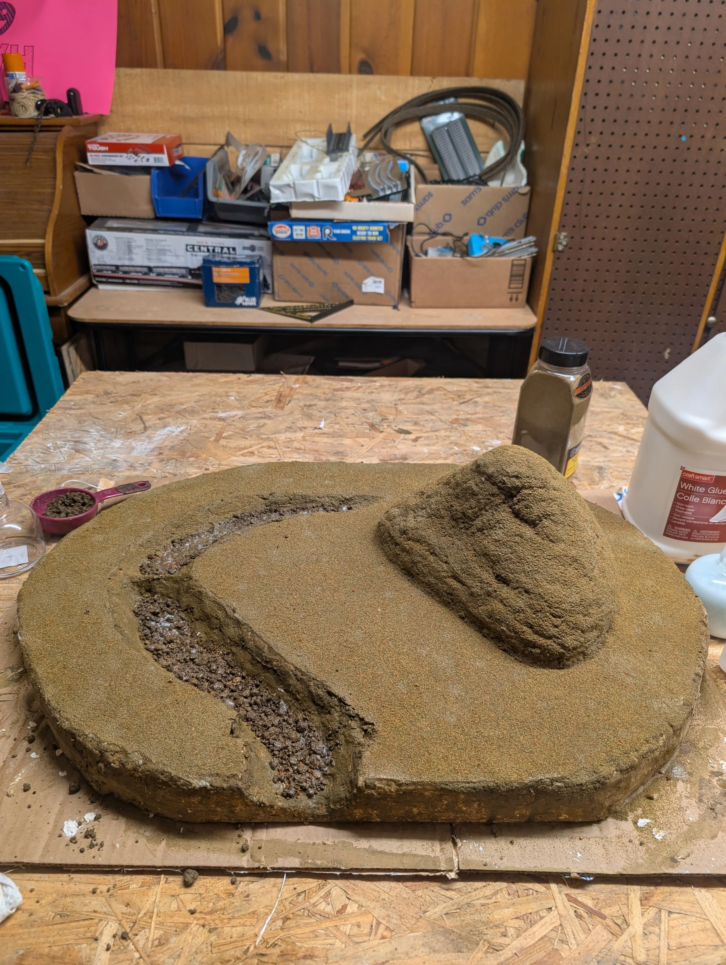 A workbench with a partial model of a terrain or landscape, featuring a curved path and a raised mound covered in a textured material, likely for a diorama or model railway.