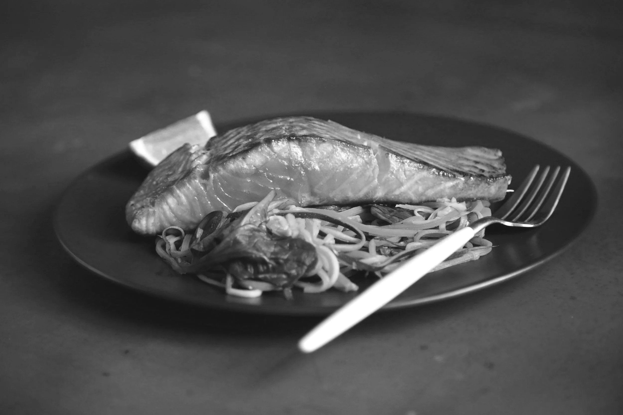 A cooked salmon fillet served on a black plate with a side of spiralized vegetables and a lime wedge, with a fork and white chopsticks placed on the plate.