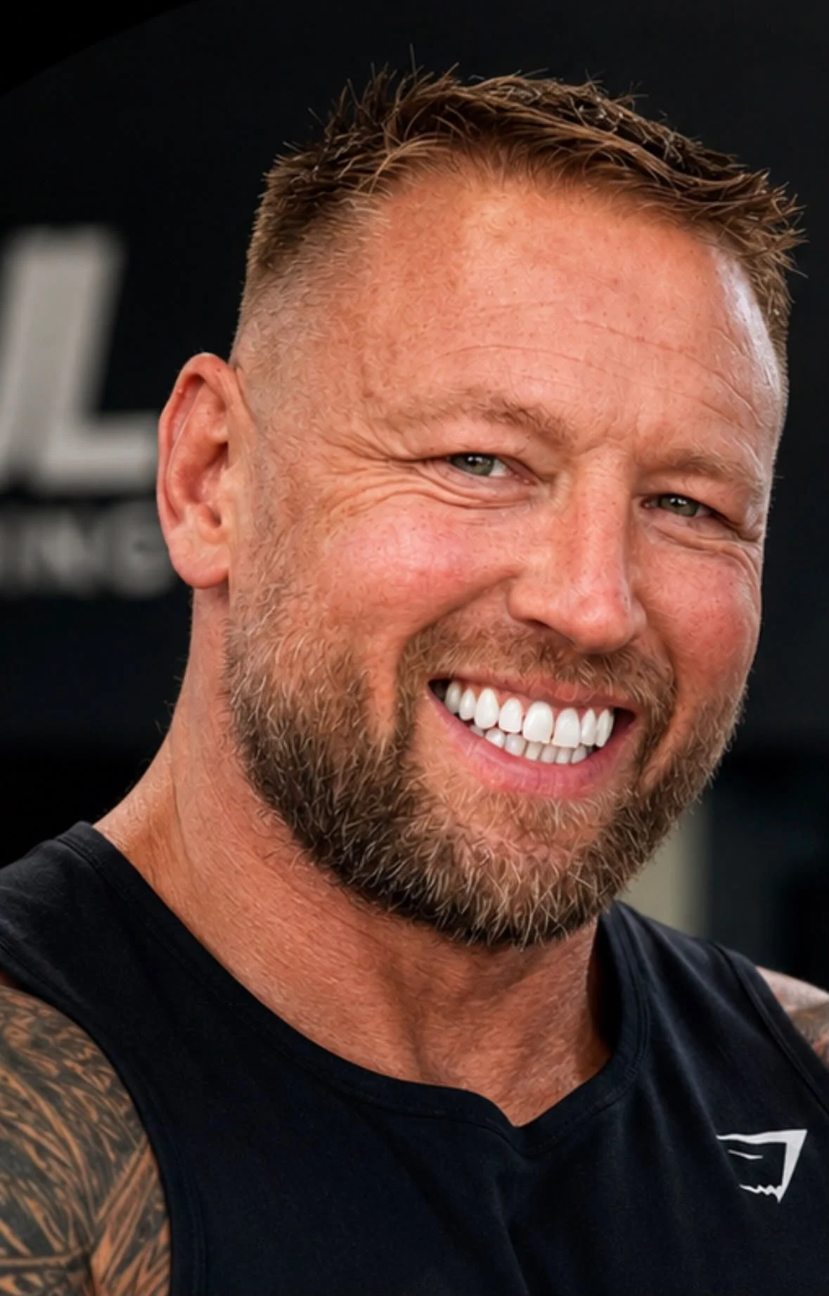 A smiling personal trainer from coventry with short hair, a beard, and tattoos on his arm, wearing a black sleeveless shirt, in an indoor setting.