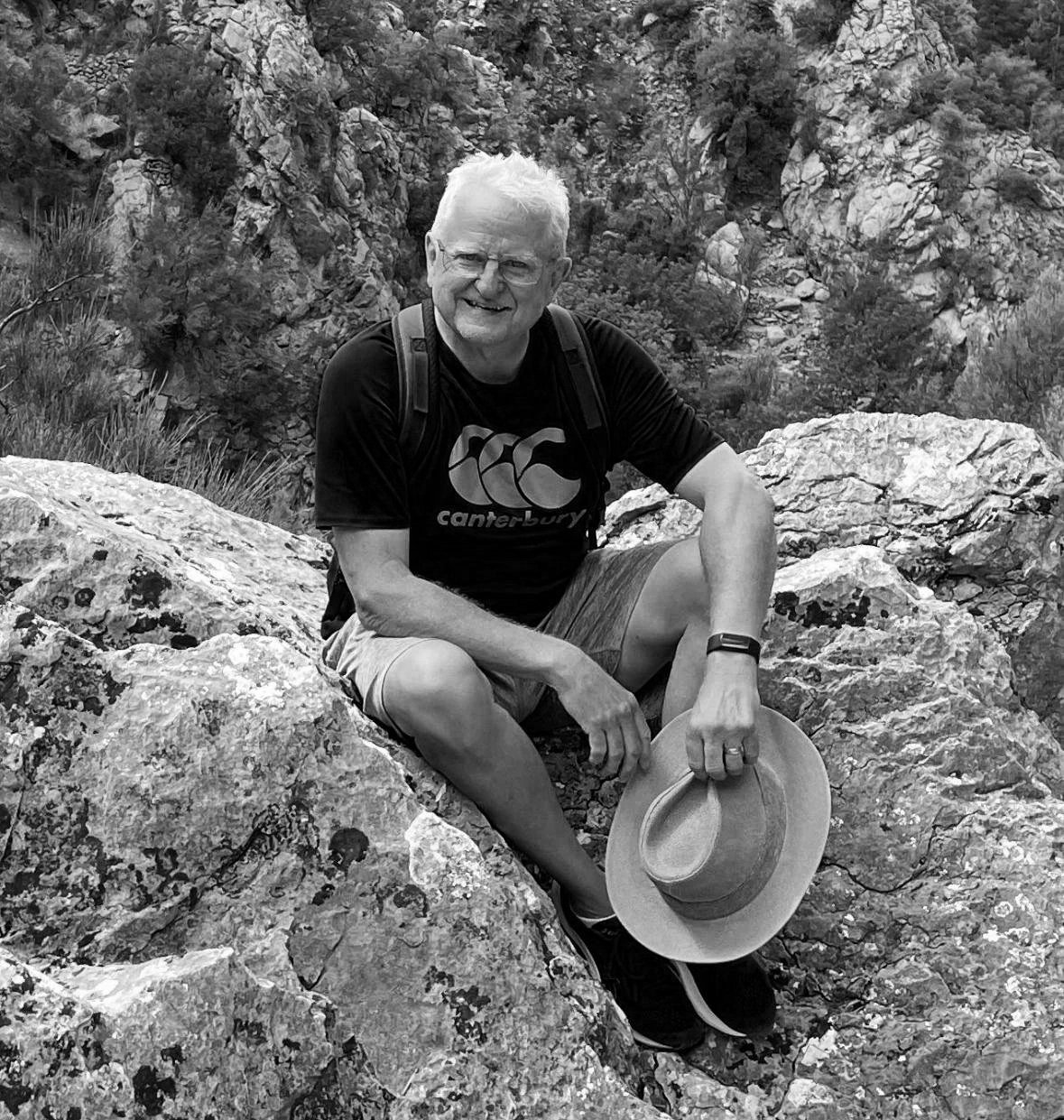 An older man with glasses and a beard, sitting on rocks in a mountainous outdoor setting, smiling, holding a hat, wearing a Canterbury t-shirt, shorts, and a backpack.