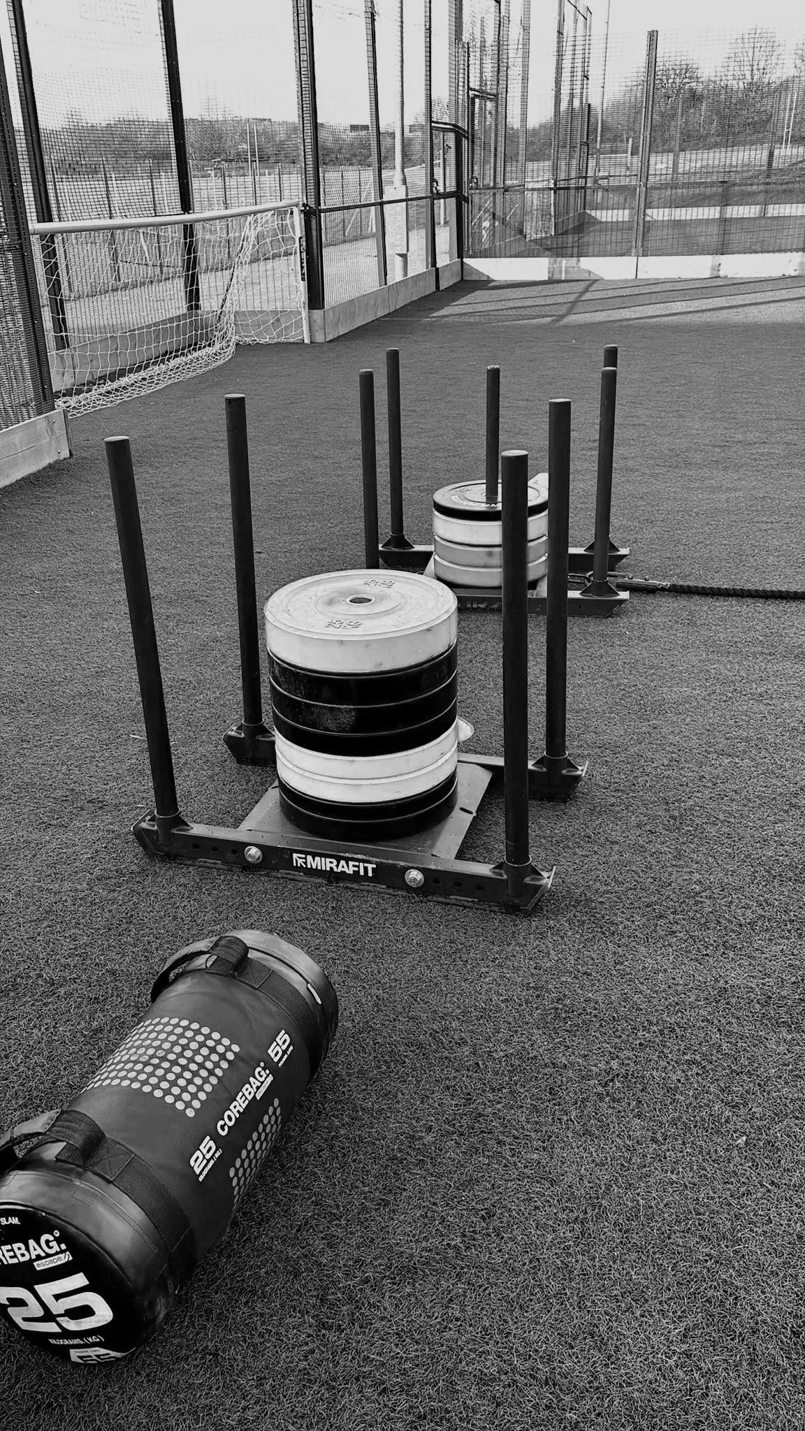 Outdoor gym area with a weighted sled, dumbbells, and artificial turf, surrounded by a fenced sports field and trees in the distance.