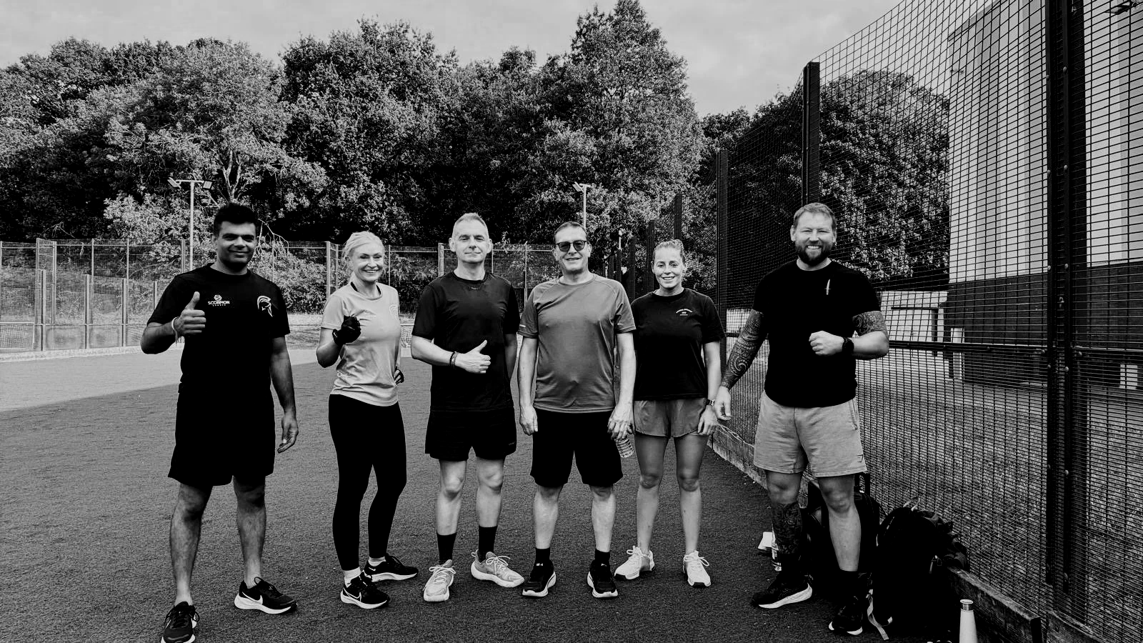 Group of six people standing on a tennis court, dressed in athletic clothing, with trees and a fence in the background.
