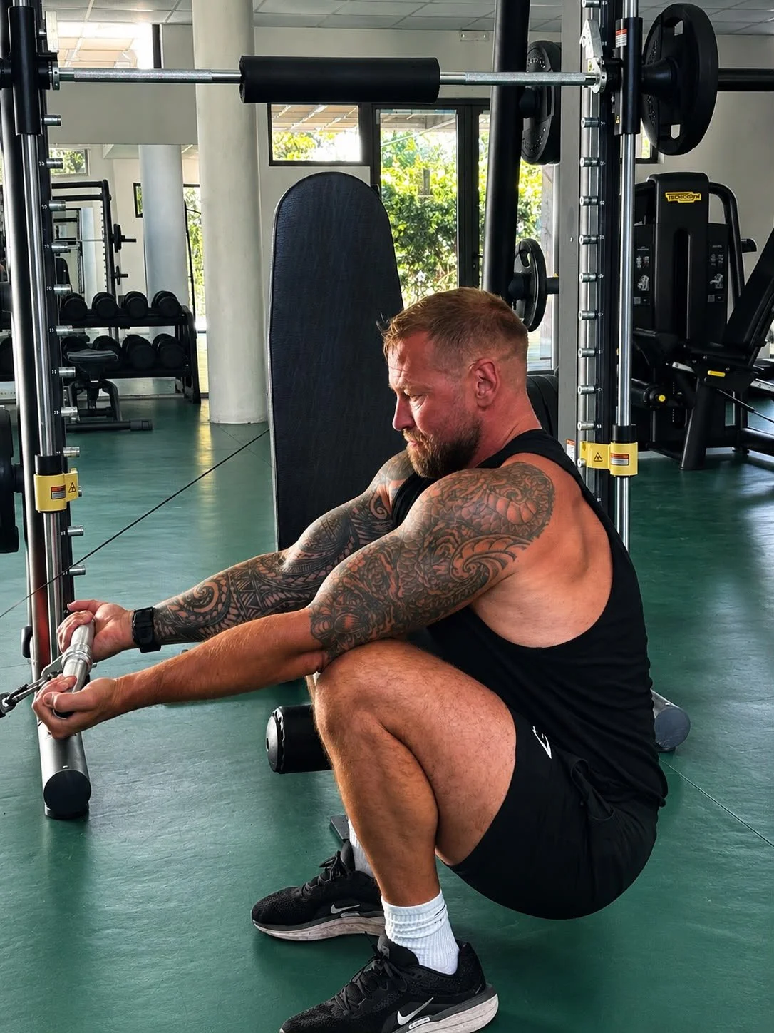 A man with tattoos on his arms and a beard, dressed in a black tank top and shorts, is performing a seated cable row exercise at a gym. He is squatting with his feet flat on the ground, holding the cable handle with both hands, with a focused expression.