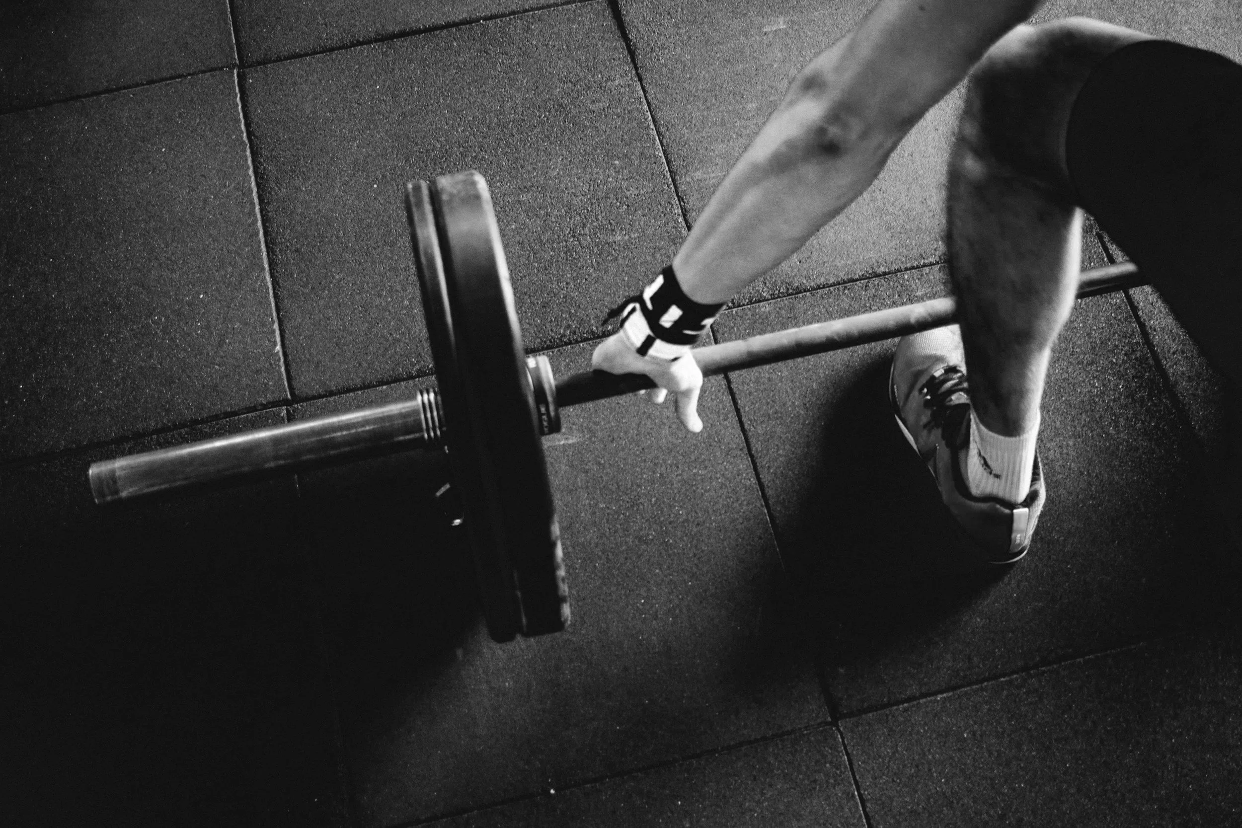 Person gripping a barbell during a workout at the gym. The focus is on the person's hand, arm, and a portion of the leg, with a rubber gym floor in the background.