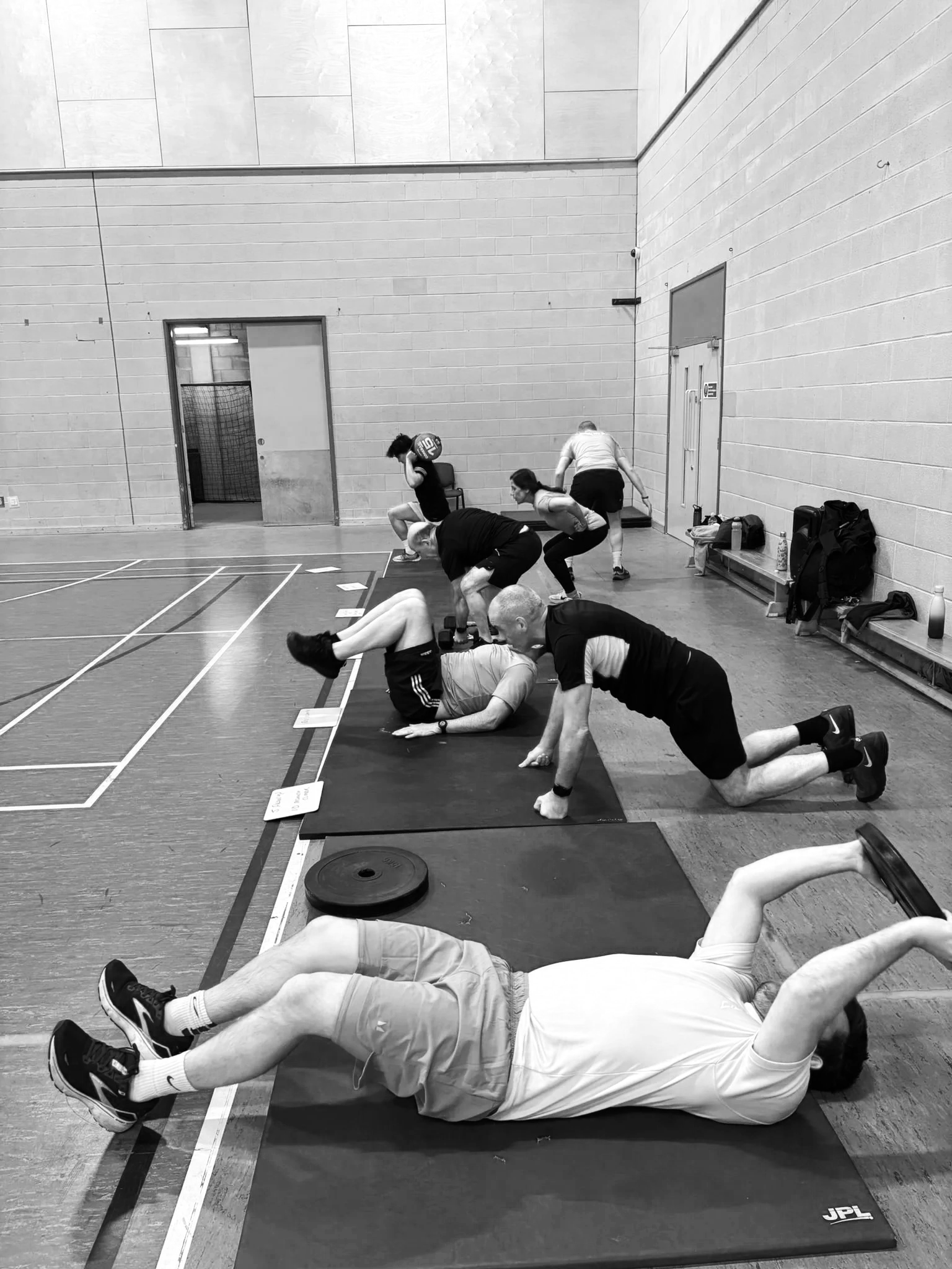 People practicing various exercises on mats in a gymnasium with cinder block walls, including planks and squats.