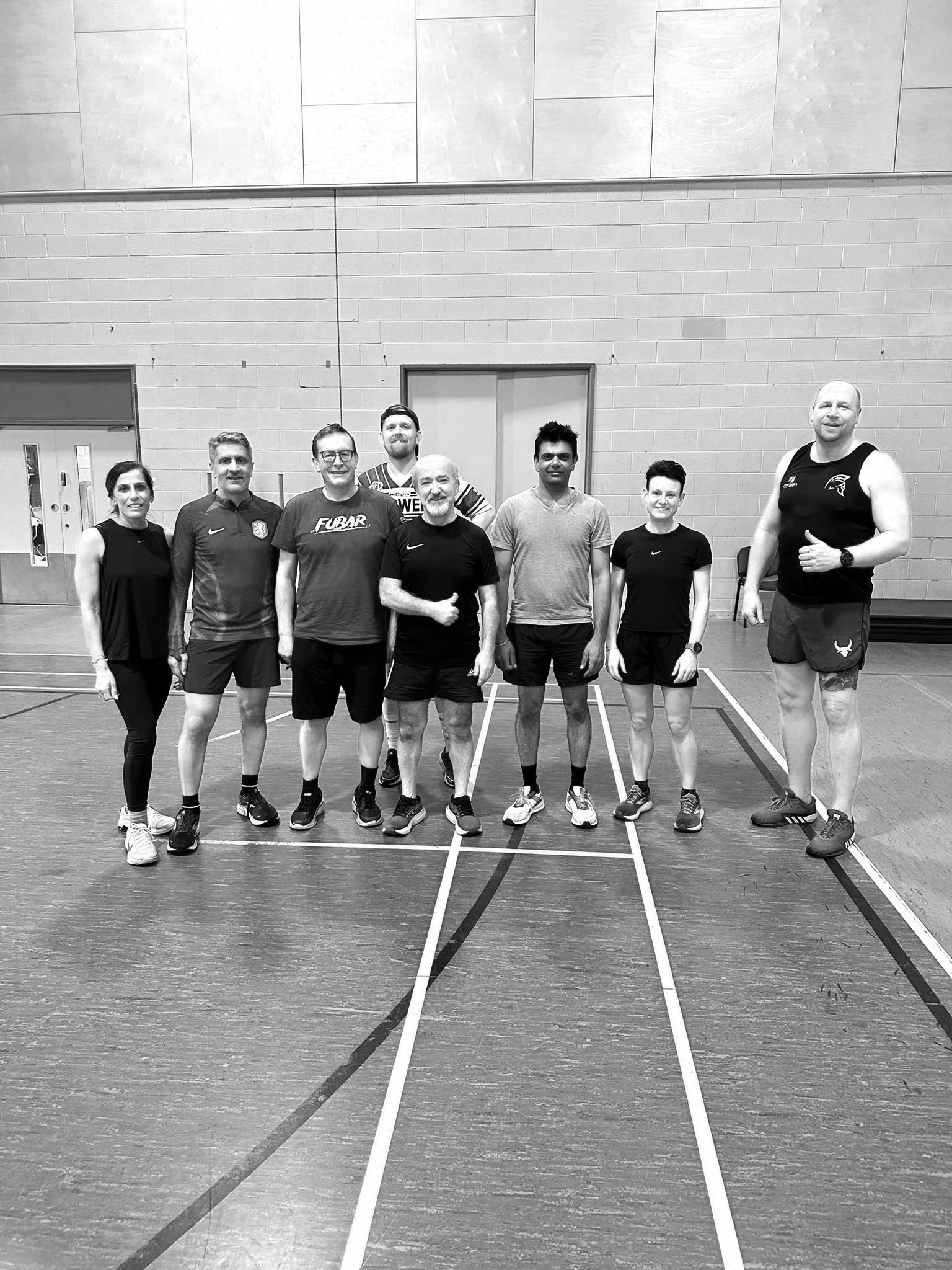 A group of nine people standing in a gymnasium, posing for a photo. They are all wearing athletic clothes and sneakers, and appear to be of diverse ages and backgrounds.