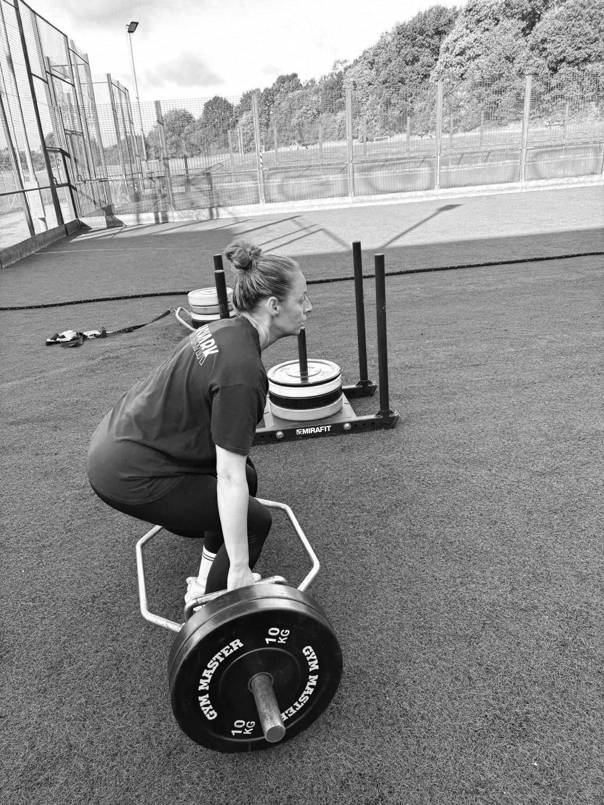 A woman in athletic clothing lifts a barbell with weights in an outdoor gym setting.
