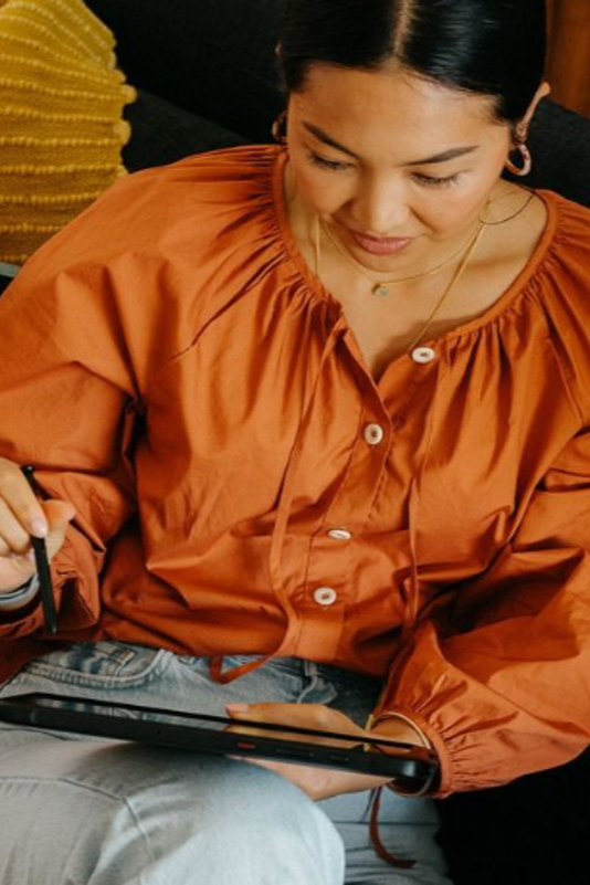Woman wearing an orange blouse sitting and writing on a digital tablet.