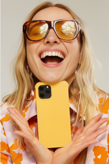 Woman with blonde hair wearing large sunglasses and a colorful shirt, taking a selfie with a yellow phone while smiling.