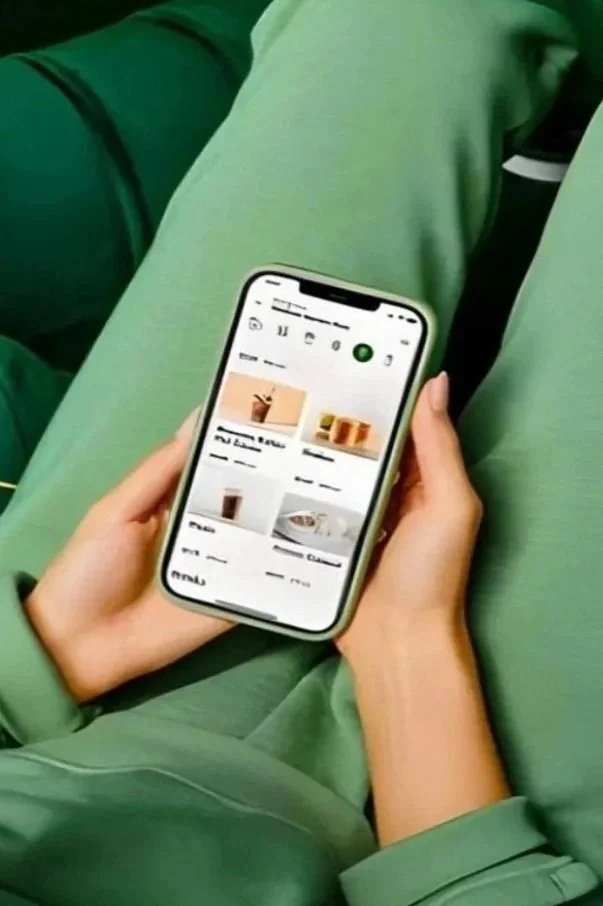 Person holding a smartphone, browsing a shopping app with food and drink options, while sitting cross-legged in green pants.