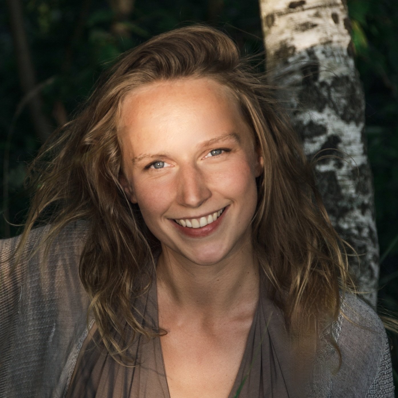 A woman with light skin, wavy blonde hair, and blue eyes smiling outdoors near a tree with a background of green foliage.