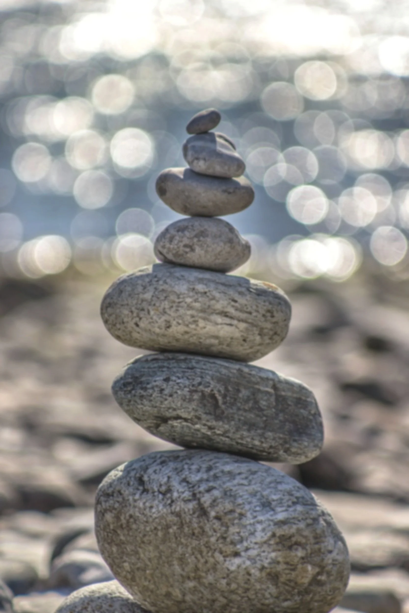 A stack of smooth, rounded stones balanced on top of each other on a beach with sunlight reflecting off the water in the background.