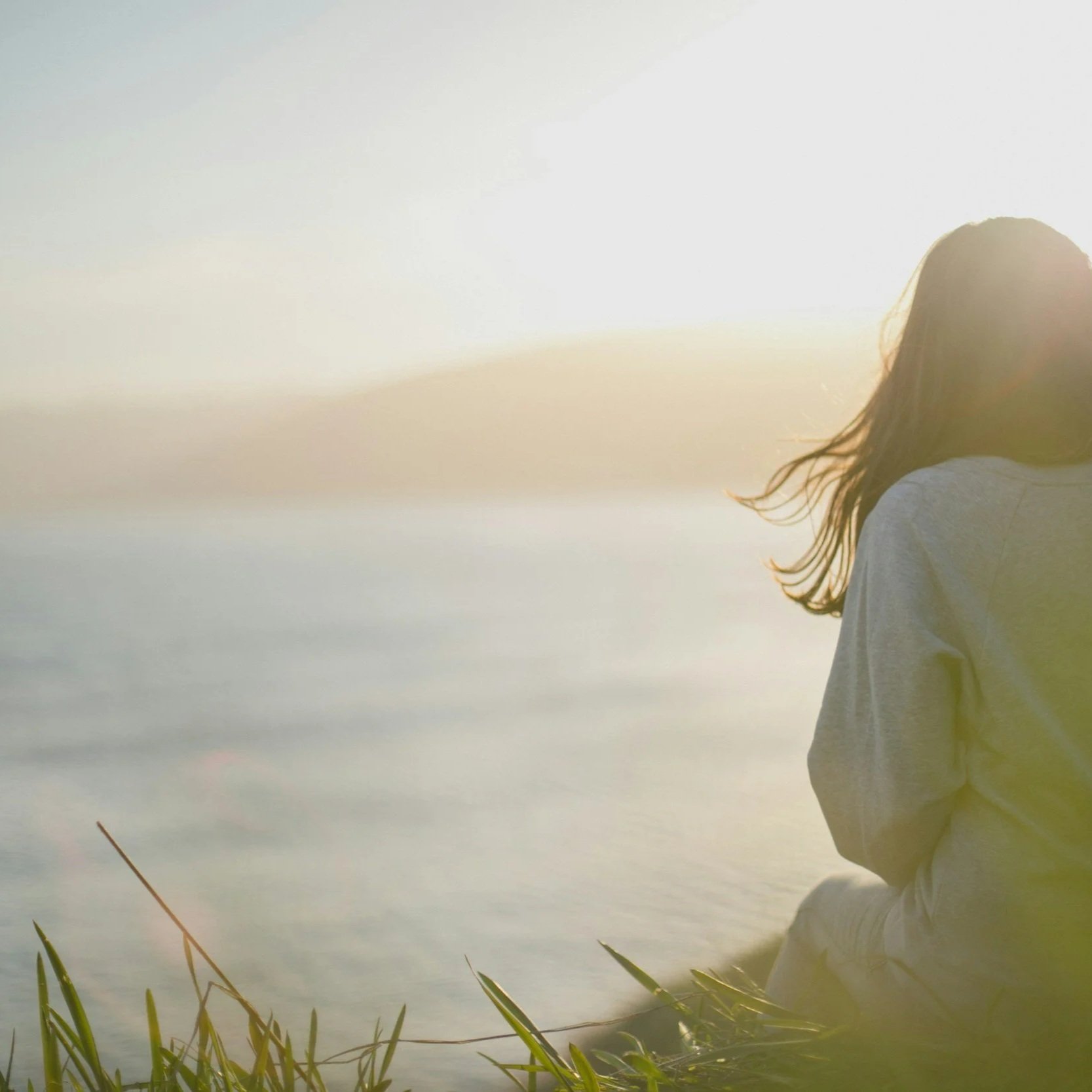 A person with long hair standing near a body of water during sunrise or sunset, wearing a white garment.