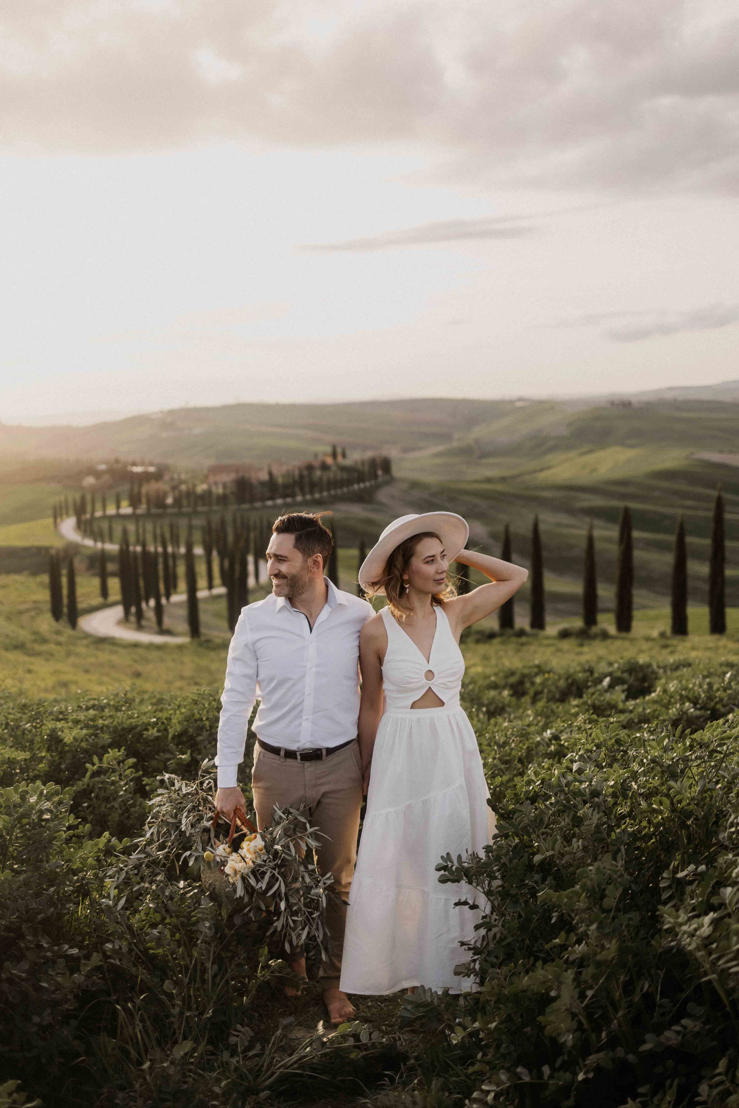 A couple walking through a vineyard with rolling hills and cypress trees in the background, during sunset, holding a bouquet of flowers.