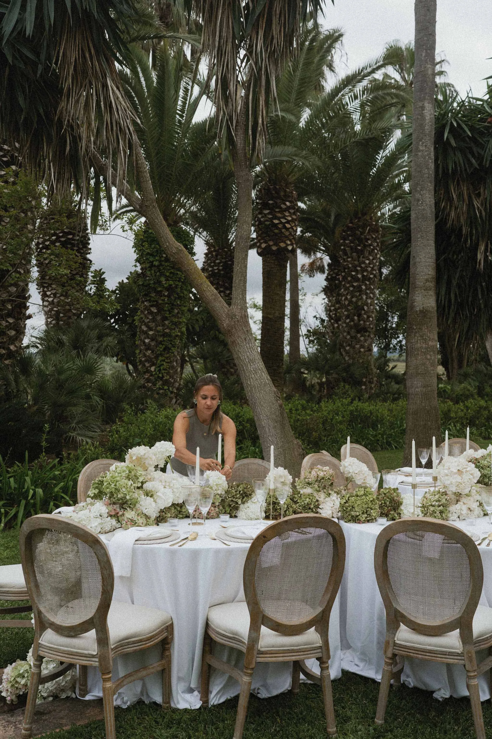 Outdoor wedding reception setup with a round table, white tablecloth, floral centerpieces, and chairs, surrounded by tall palm trees and lush greenery.