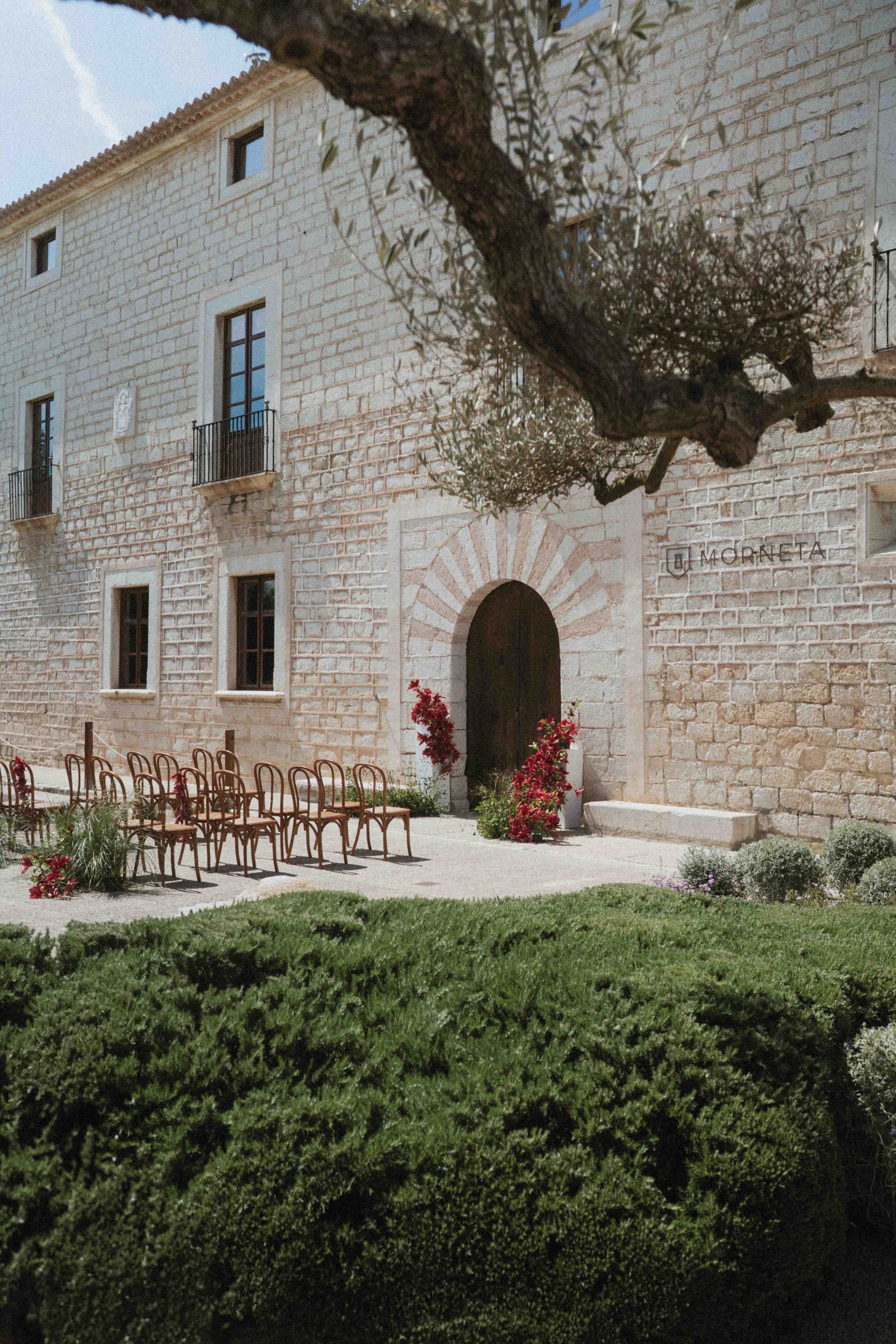 A stone building with multiple windows and small balconies. The words 'MORNETA' are on the wall beside the entrance. In front, there are several rows of empty chairs and plants with red flowers, a small pathway, and a green hedge.