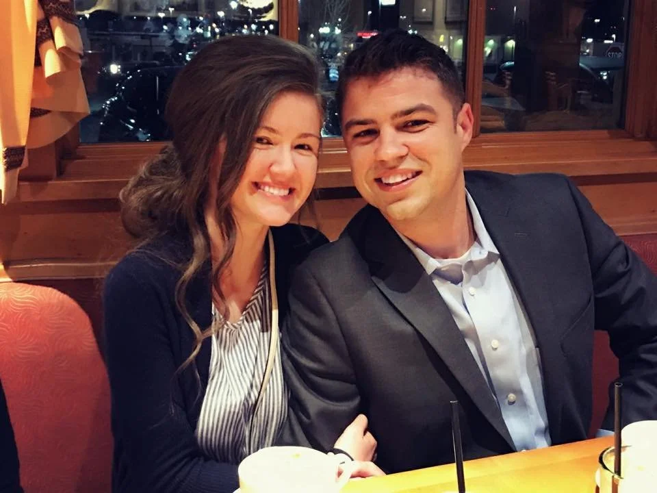 A smiling young woman (Erika) with long, wavy brown hair and a young man - Nathanael Schultz - with short, dark hair sitting together at a restaurant table. Both are dressed nicely.