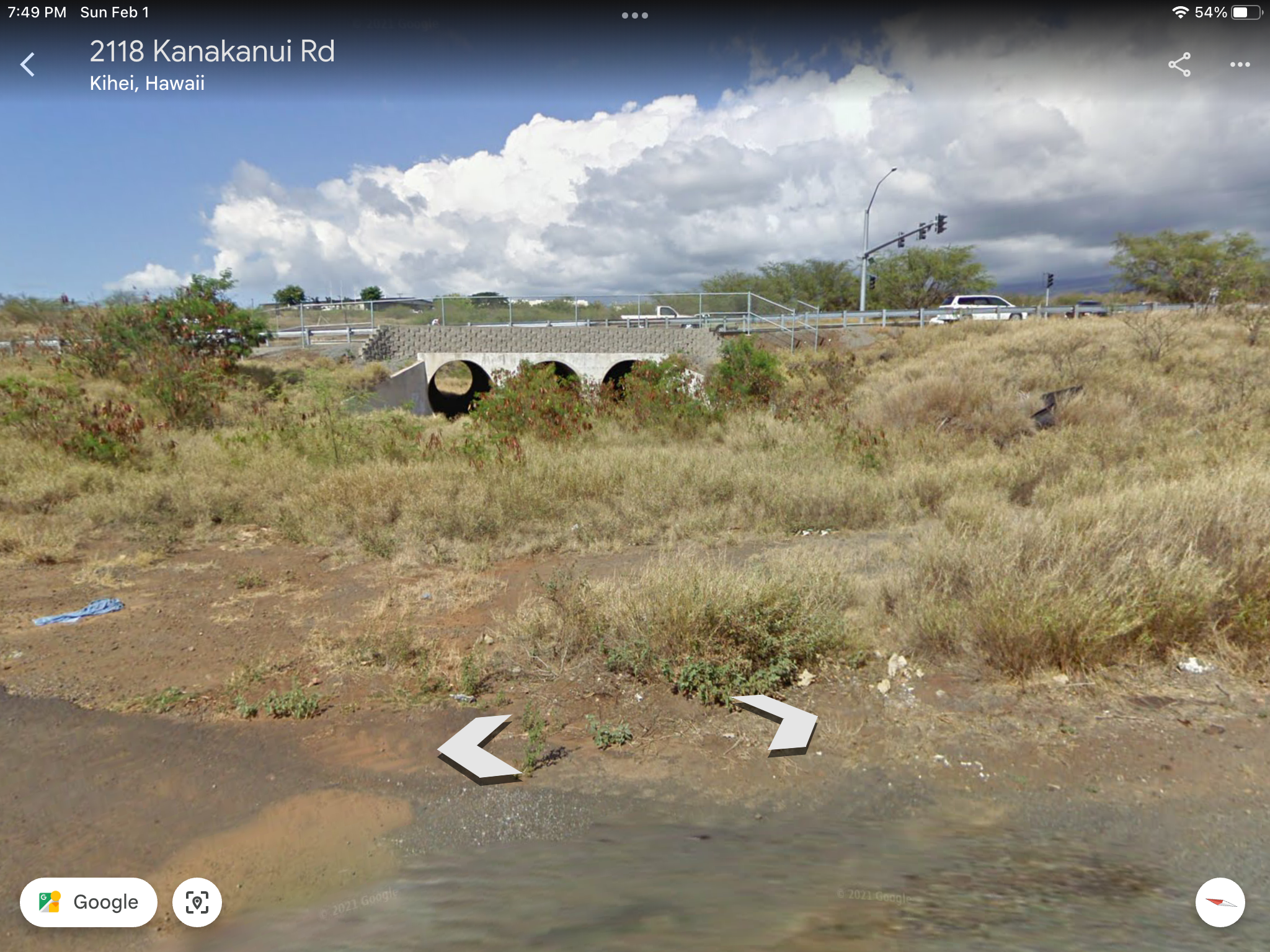 A rural scene with a bridge over a dry grassy area, with a street and traffic lights in the background. There are a few scattered bushes and a small amount of litter in the foreground.