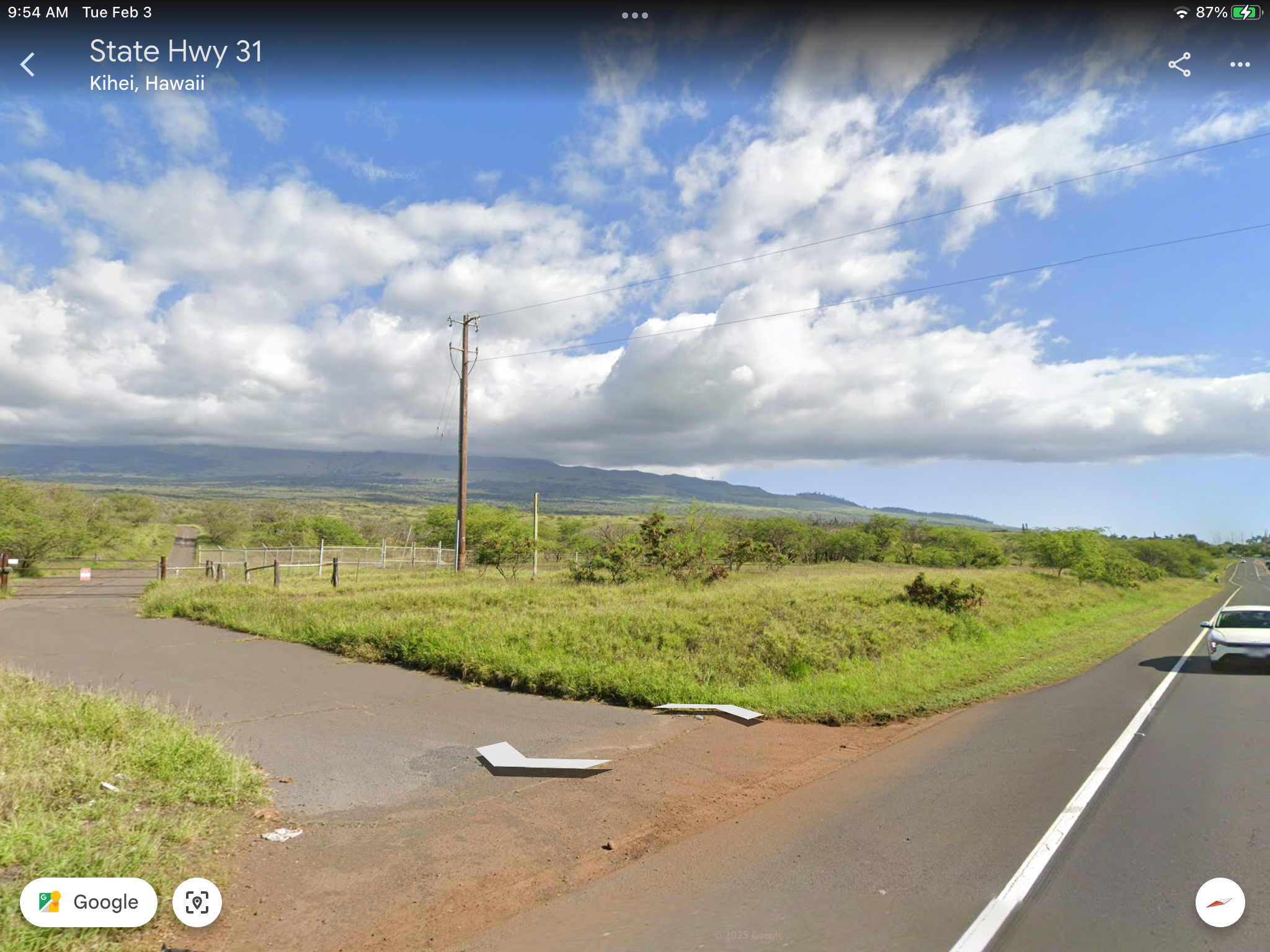View of a rural highway scene in Kihei, Hawaii. The image shows a two-lane road with cars, grassy fields, a utility pole, and a fenced area. A mountain range is visible in the distance under a partly cloudy sky.