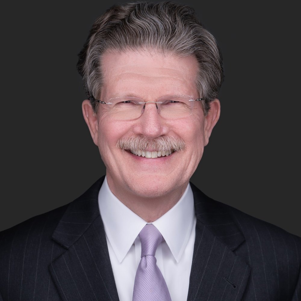 Portrait of Garry Stanberry wearing glasses, a dark suit, white shirt, and light purple tie, smiling against a dark background.