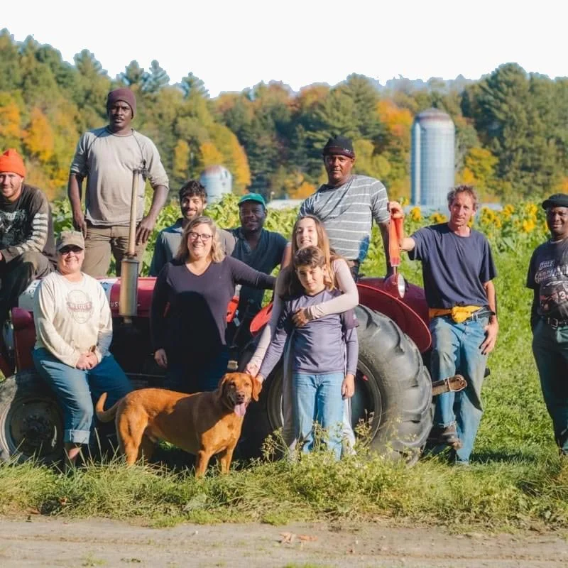 The crew from Foote Brook Farm and a dog gathered in front of a tractor on the farm. The background features green fields, trees, and farm silos, suggesting an autumn setting.