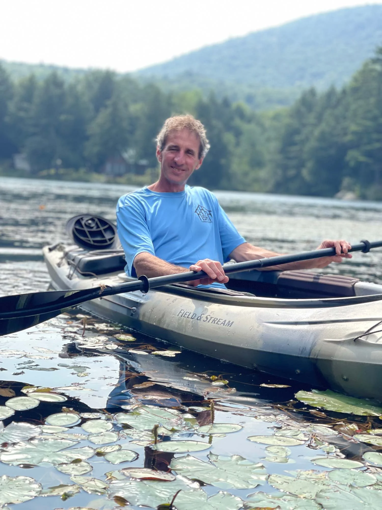 A man paddling a kayak on a lake with lily pads, surrounded by trees and mountains in the background.