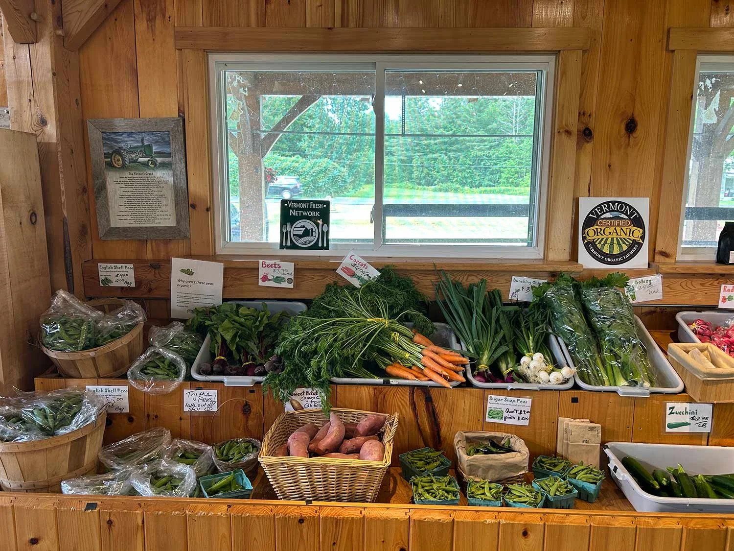 Display of fresh farm vegetables including carrots, onions, beets, zucchinis, and snap peas inside a wooden farm stand with signs for Vermont Organic and Vermont Fresh Network.