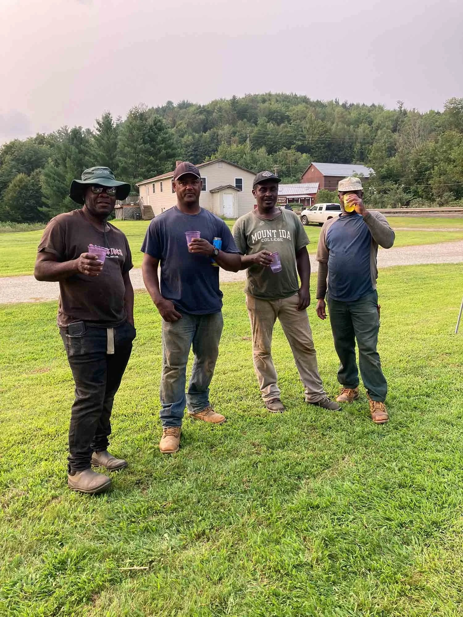 Four men standing on a grassy field, holding drinks, with trees, houses, and a car in the background.