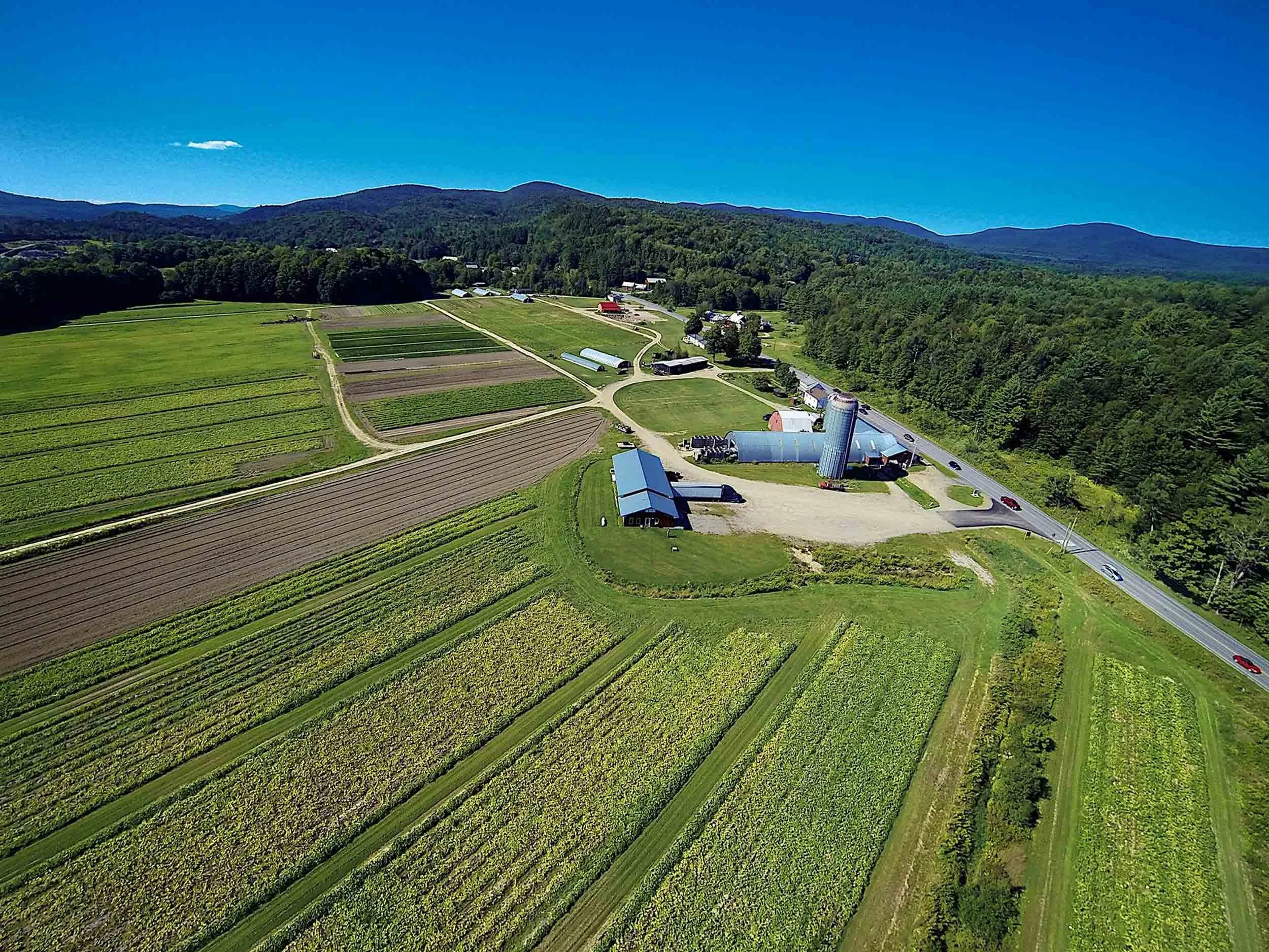 Aerial view of a farm with fields, barns, silos, and a main road, surrounded by green hills and forest under a clear blue sky.