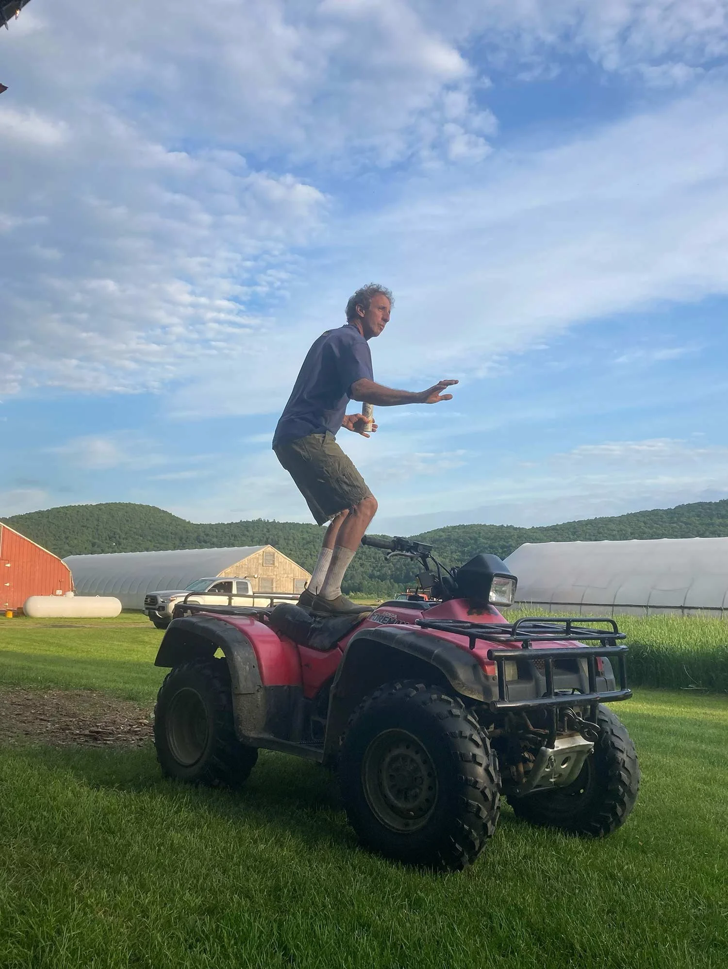 A man standing on top of a red  ATV, balancing with arms outstretched, in a rural field with farm buildings and green hills in the background.