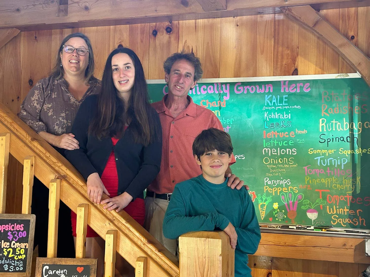 A group of four people posing for a photo in front of a colorful vegetable and produce sign. The group includes two women, a man, and a boy, standing on a wooden staircase inside a rustic wooden building.