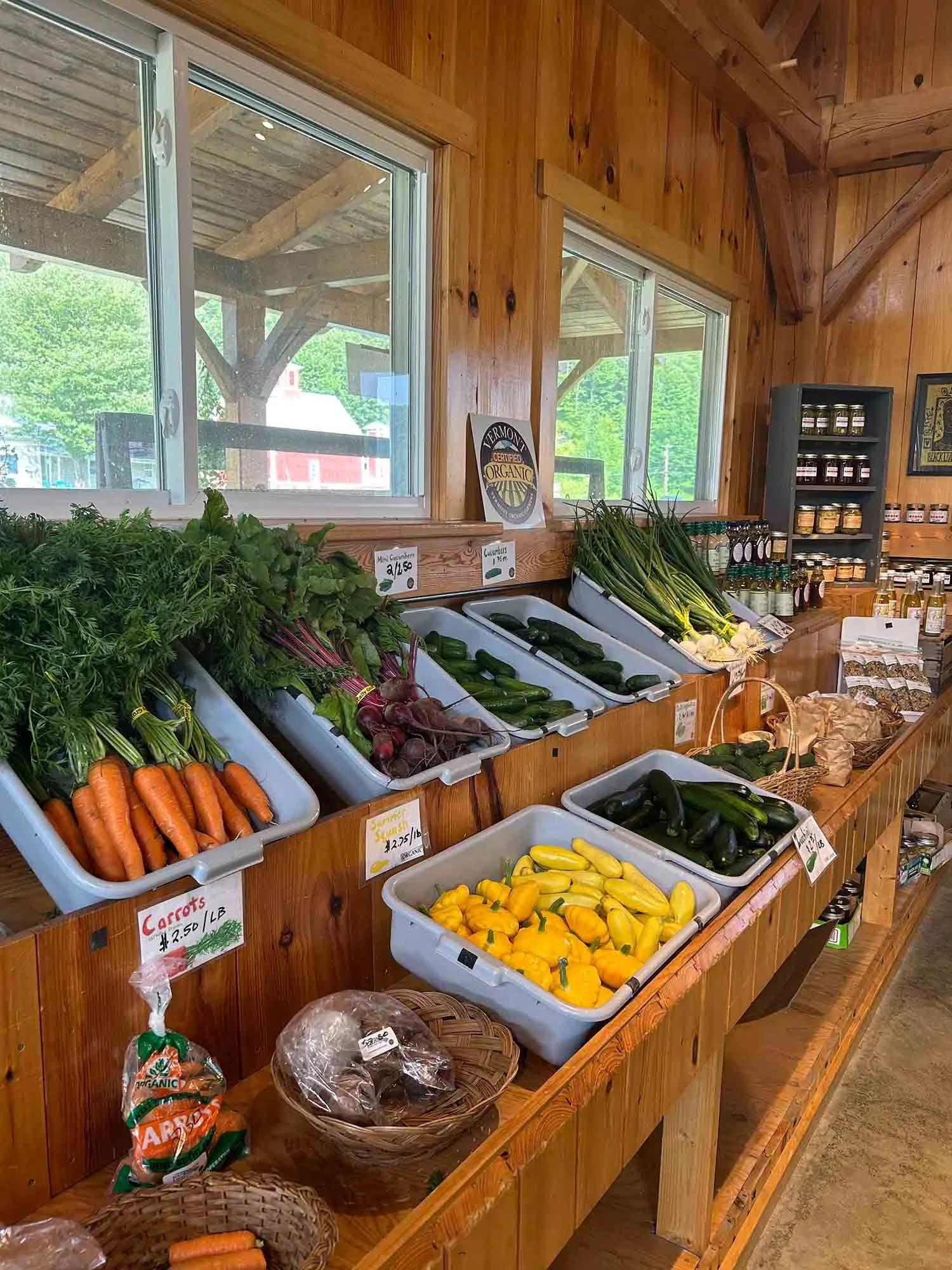 Fresh vegetables, including carrots, beets, zucchini, leeks, and yellow squash, are displayed in bins inside the Foote Brook Farm Store with large windows showing an outdoor wooden patio and green trees.