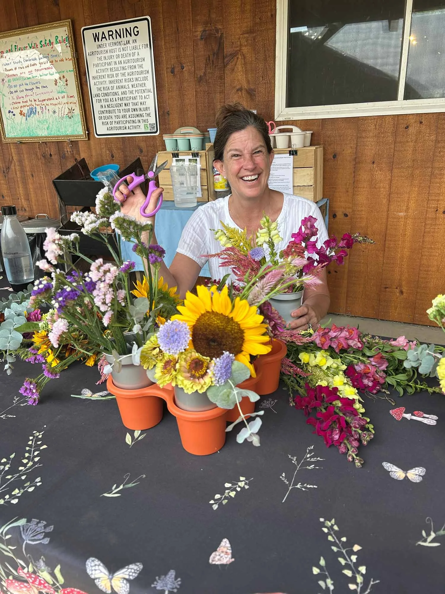 A smiling woman arranging a bouquet of colorful flowers at a flower arranging table, with various floral arrangements and craft supplies on the table, and a wooden wall and window in the background.