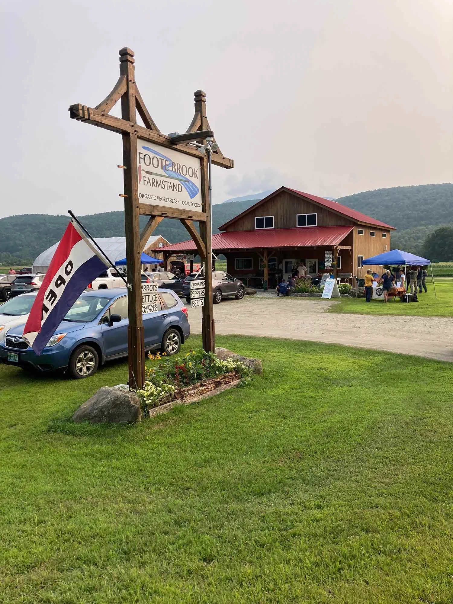 Foot Brook Farmstand sign and farm store with mountain view in the background, nearby cars, and people under tents.