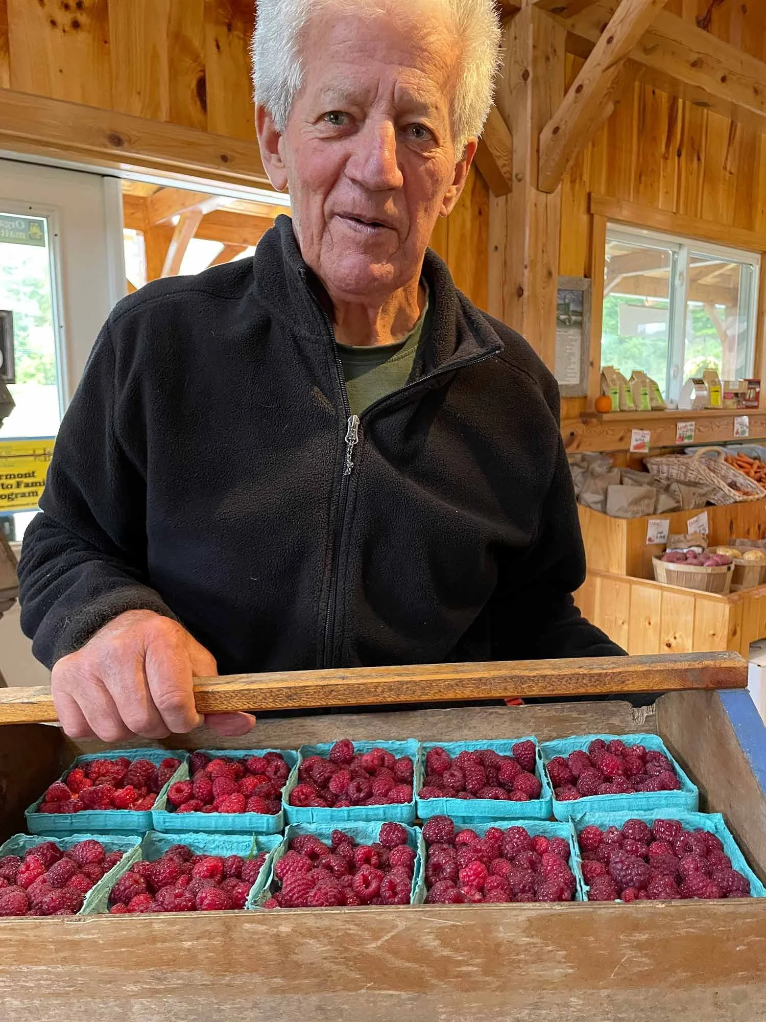 An man in a black fleece jacket standing behind a wooden display containing small baskets of red raspberries inside the farm stand, with a window and shelves of produce in the background.