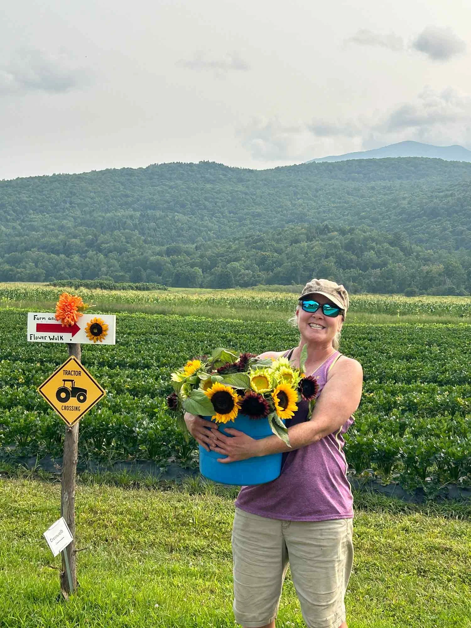 Smiling woman wearing sunglasses, holding a blue basket filled with sunflowers and dark red flowers, standing in a field of green crops with mountains in the background.