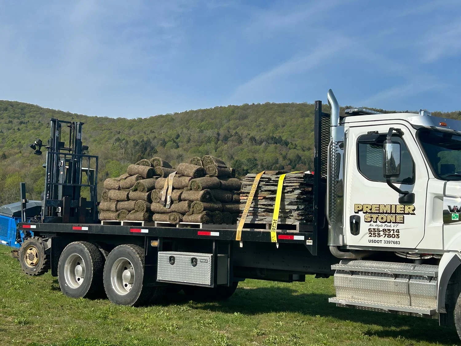 A white flatbed truck loaded with rolled sod grass parked on green grass with a hill and blue sky in background.