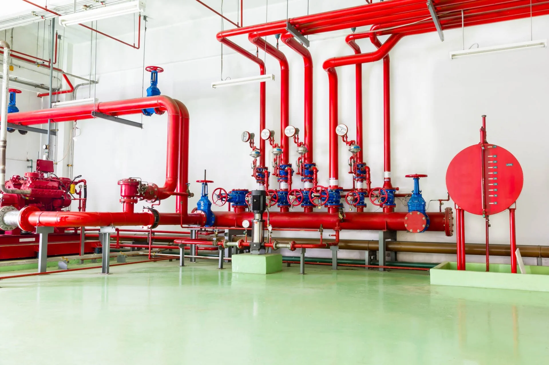 Sistema de encanamento industrial com tubos vermelhos, válvulas azuis e medidores de pressão azuis, em ambiente limpo com piso verde e paredes brancas.