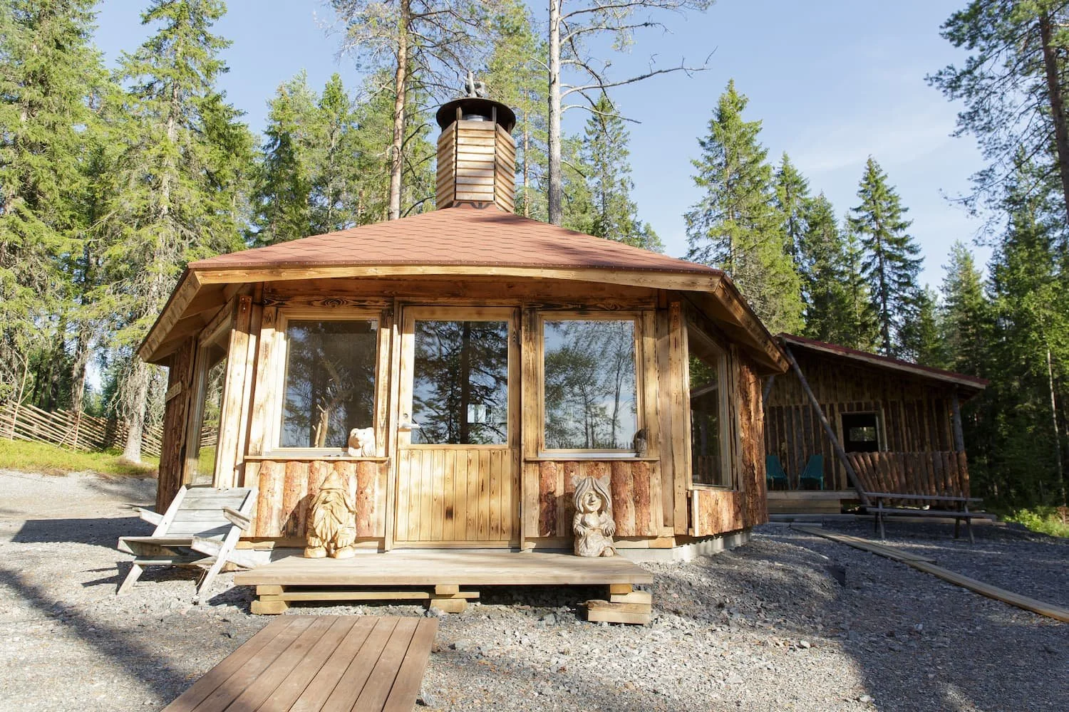 A small round wooden cabin with a brown shingled roof, situated in a forested area with tall green trees. The cabin has large windows with wooden frames, a small porch with wooden steps, and decorative wooden sculptures of bears near the entrance. There is a gravel pathway leading to the porch and a wooden walkway extending from it. In the background, there is a separate small wooden structure with an outdoor seating area. The scene is well-lit with natural sunlight.