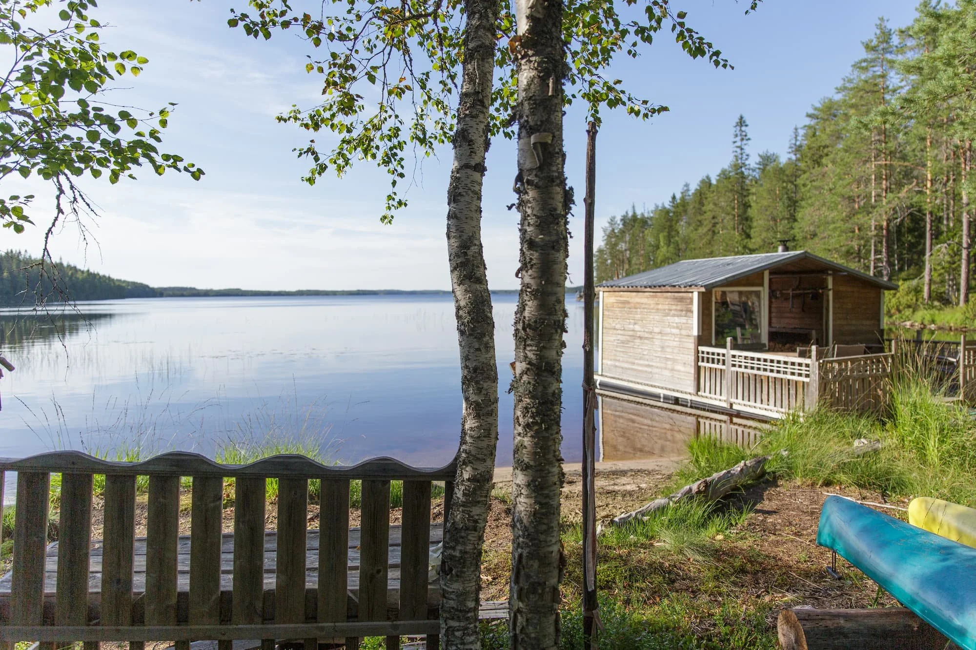 A lakeside view with two birch trees in the foreground, a small wooden house on the water's edge, a blue boat on the shore, and a background of forested land and a calm lake under a partly cloudy sky.