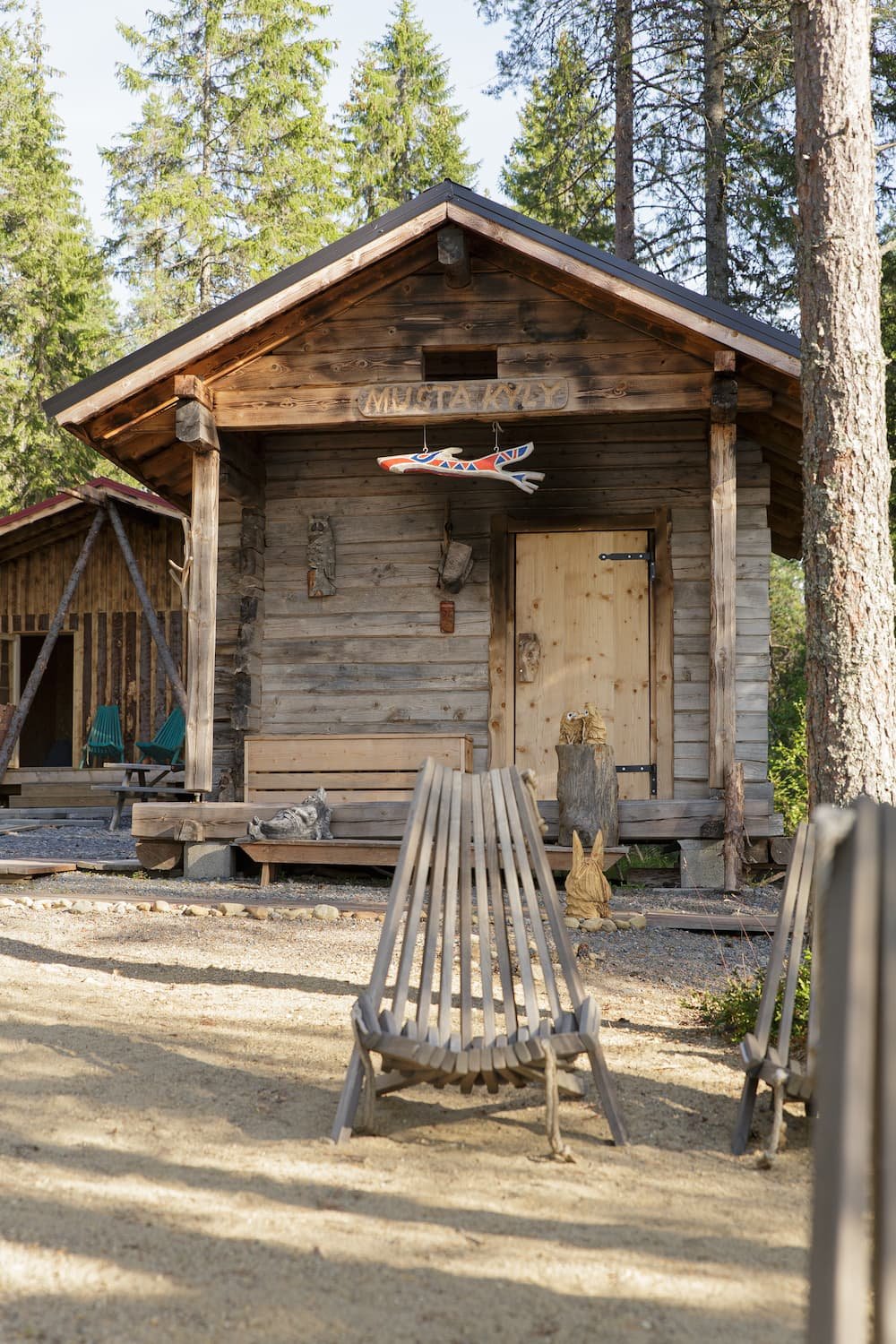 Rustic wooden cabin with a sign in Cyrillic, an interior carved wooden fish hanging from the porch ceiling, and outdoor furniture in a forested setting.