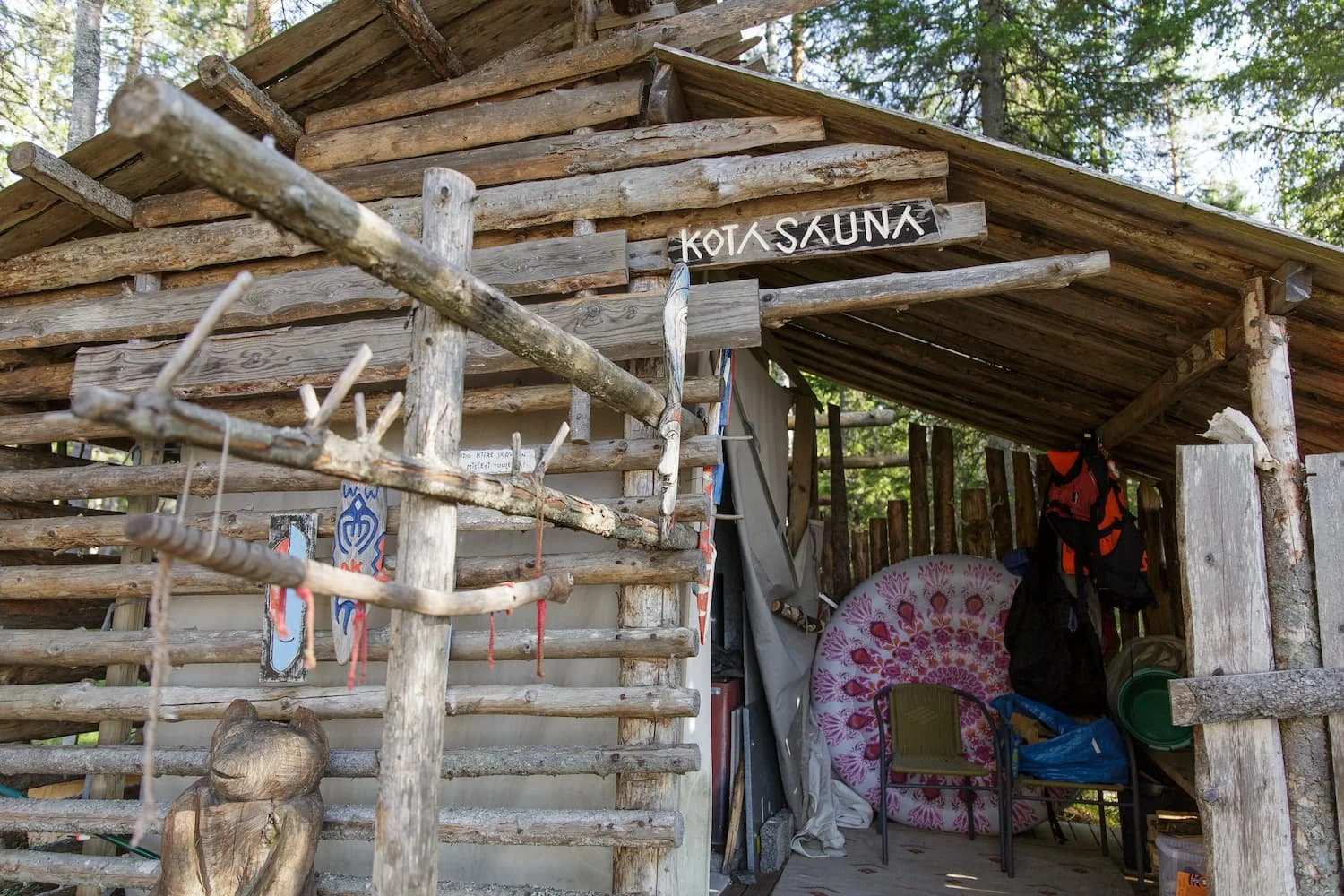 A rustic wooden shelter with a sign that reads "KOTA SAUNA". The shelter is built with rough logs and contains camping gear, chairs, and a decorative pink and white circular item, with a forest in the background.