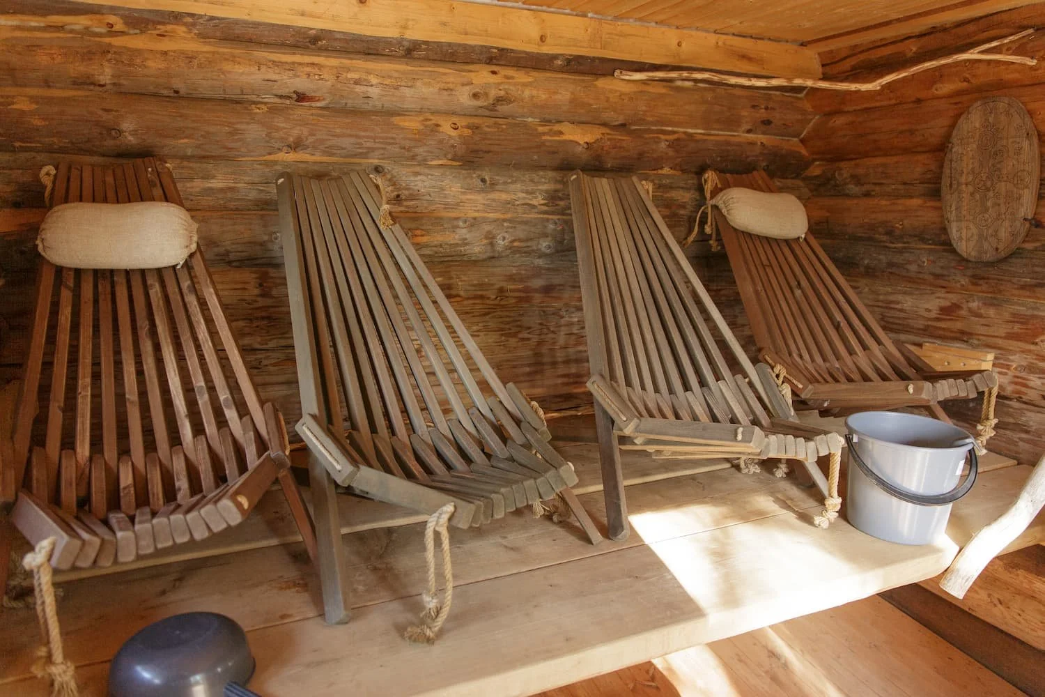 Four wooden lounge chairs with cushions inside a wood-paneled cabin, with a plastic bucket and a small wooden tray on the side.