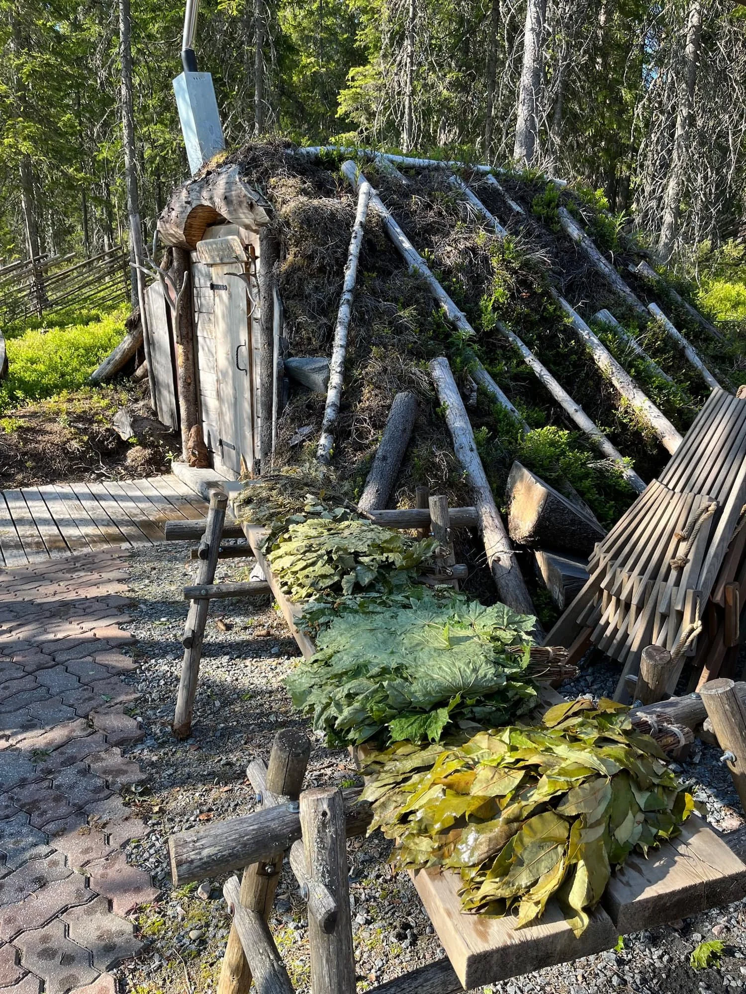 A rustic outdoor scene with a small wooden door embedded in a mound of dirt and logs, surrounded by plants, trees, and wooden furniture, with a fence in the background.